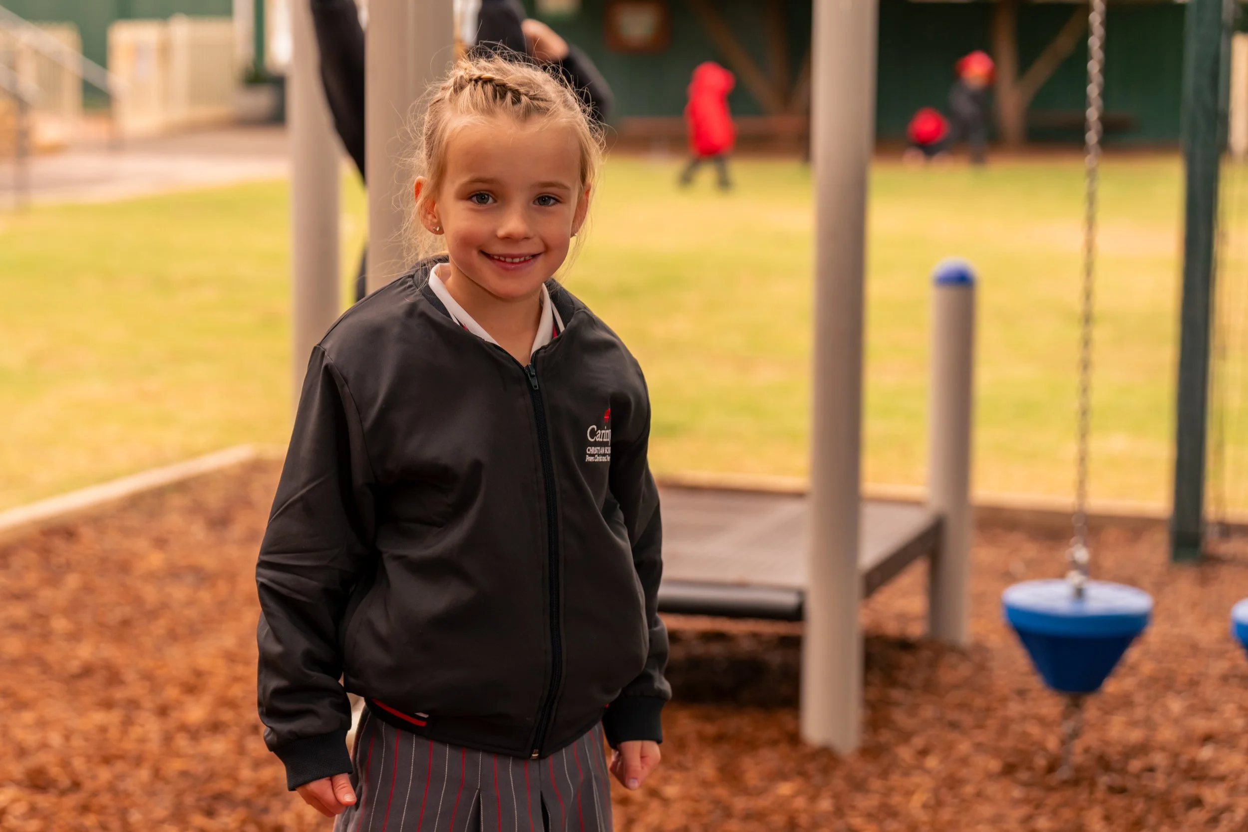 Smiling girl in black jacket and striped skirt at playground with swings and children in red jackets in the background.