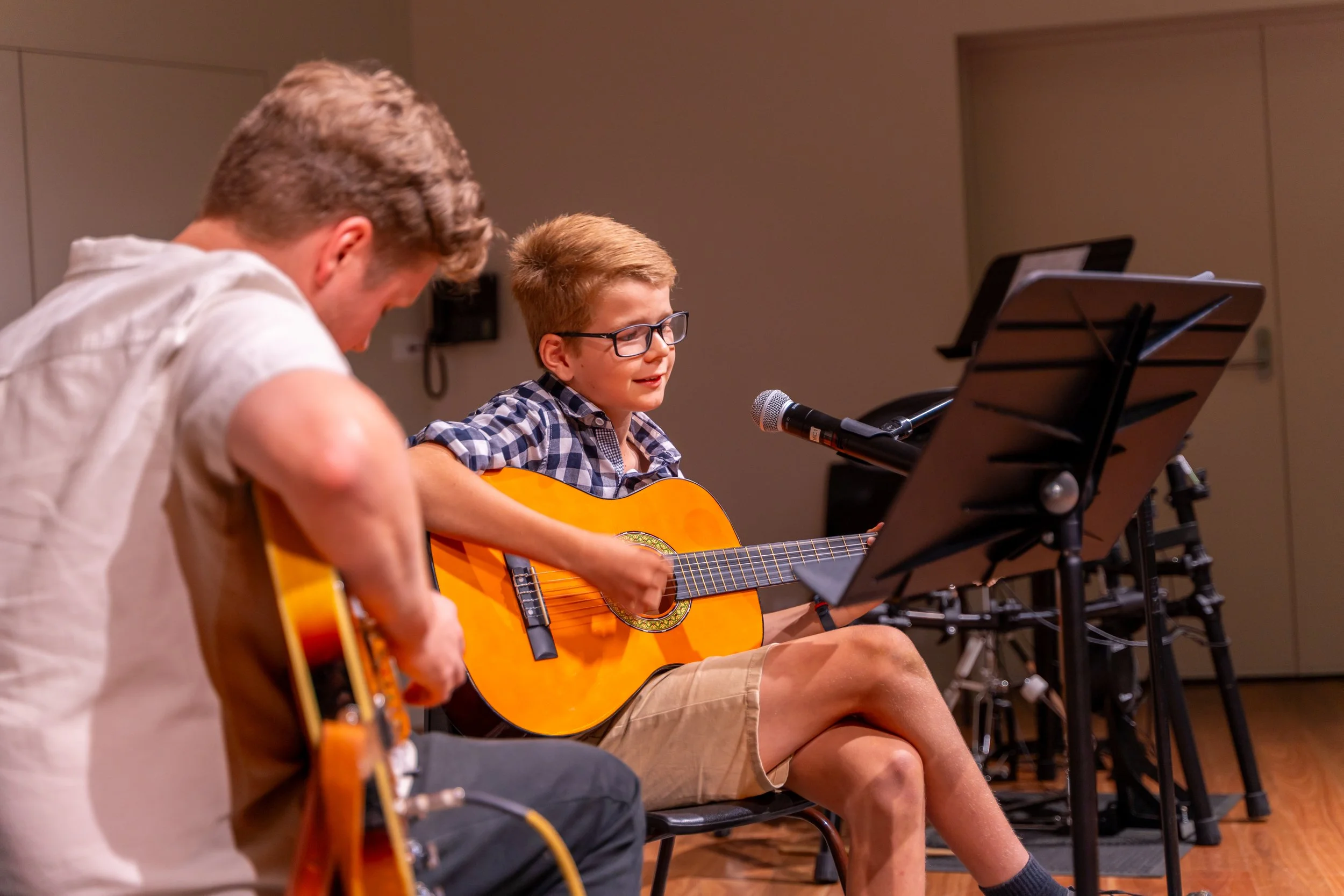Two males, one in a plaid shirt and glasses, playing an acoustic guitar and singing into a microphone, with another male, in a white shirt, playing an electric guitar next to him.