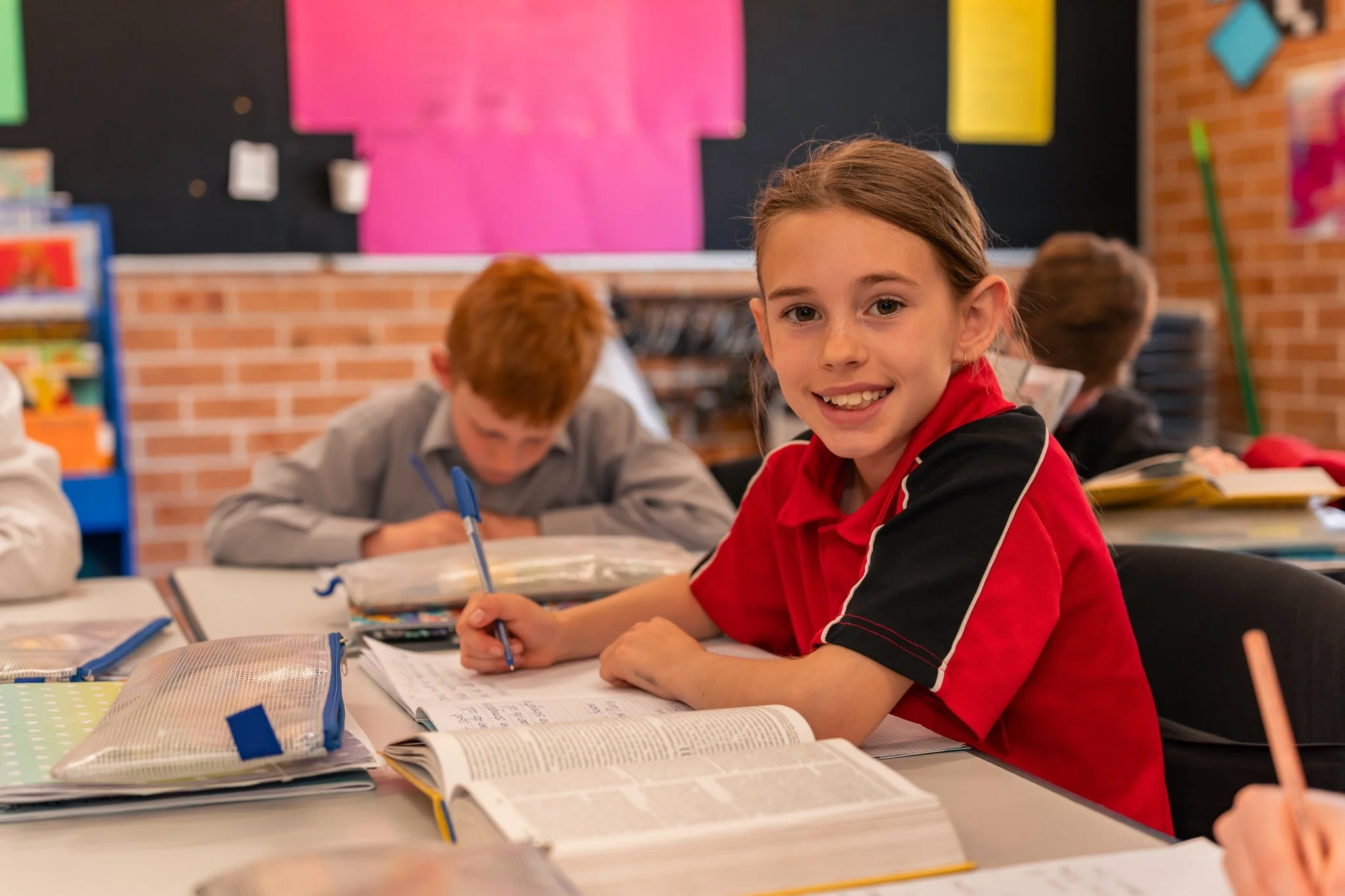 A smiling girl in a red and black school uniform sitting at a desk with open books and writing utensils in a classroom, with other students focused on their work in the background.