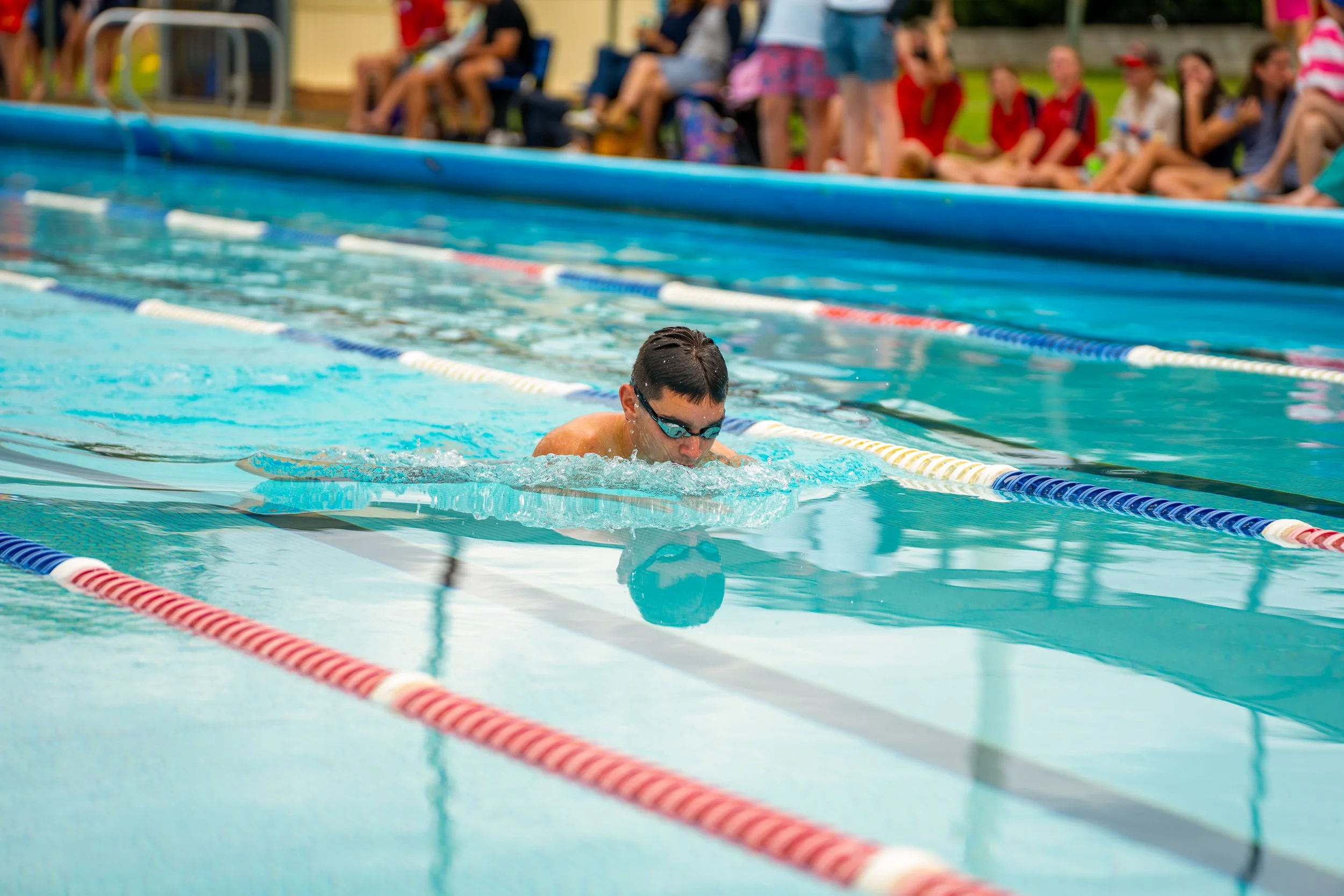 A boy with goggles swimming in an outdoor pool during a competition or practice, with spectators sitting and watching in the background.