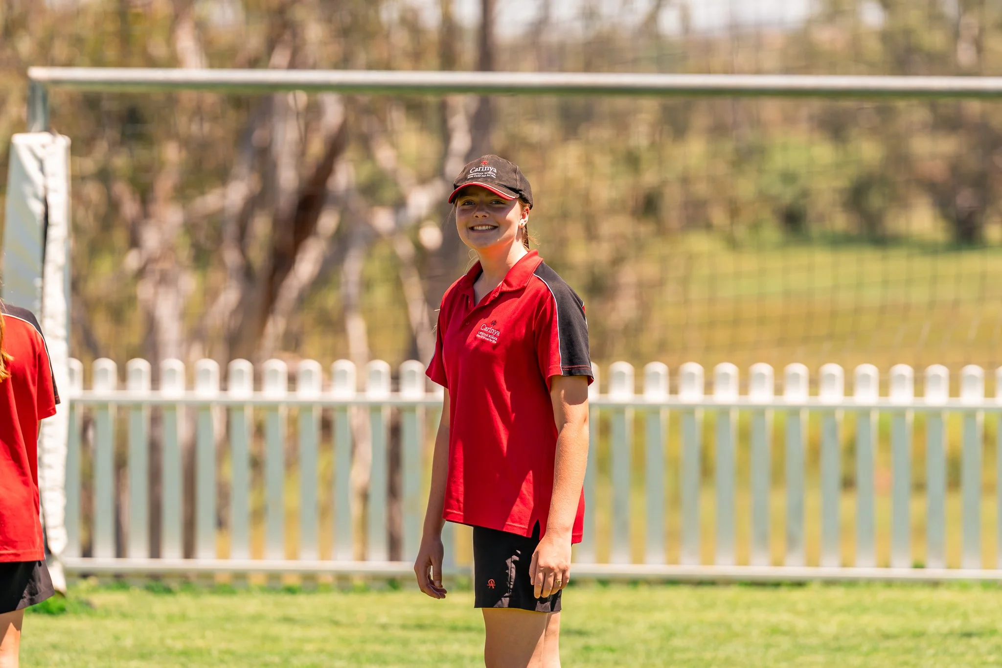 Carinya Student wearing a red sports shirt and black shorts standing on a sports field with a smile, in front of a goalpost and white picket fence at Carinya Christian School, Tamworth.