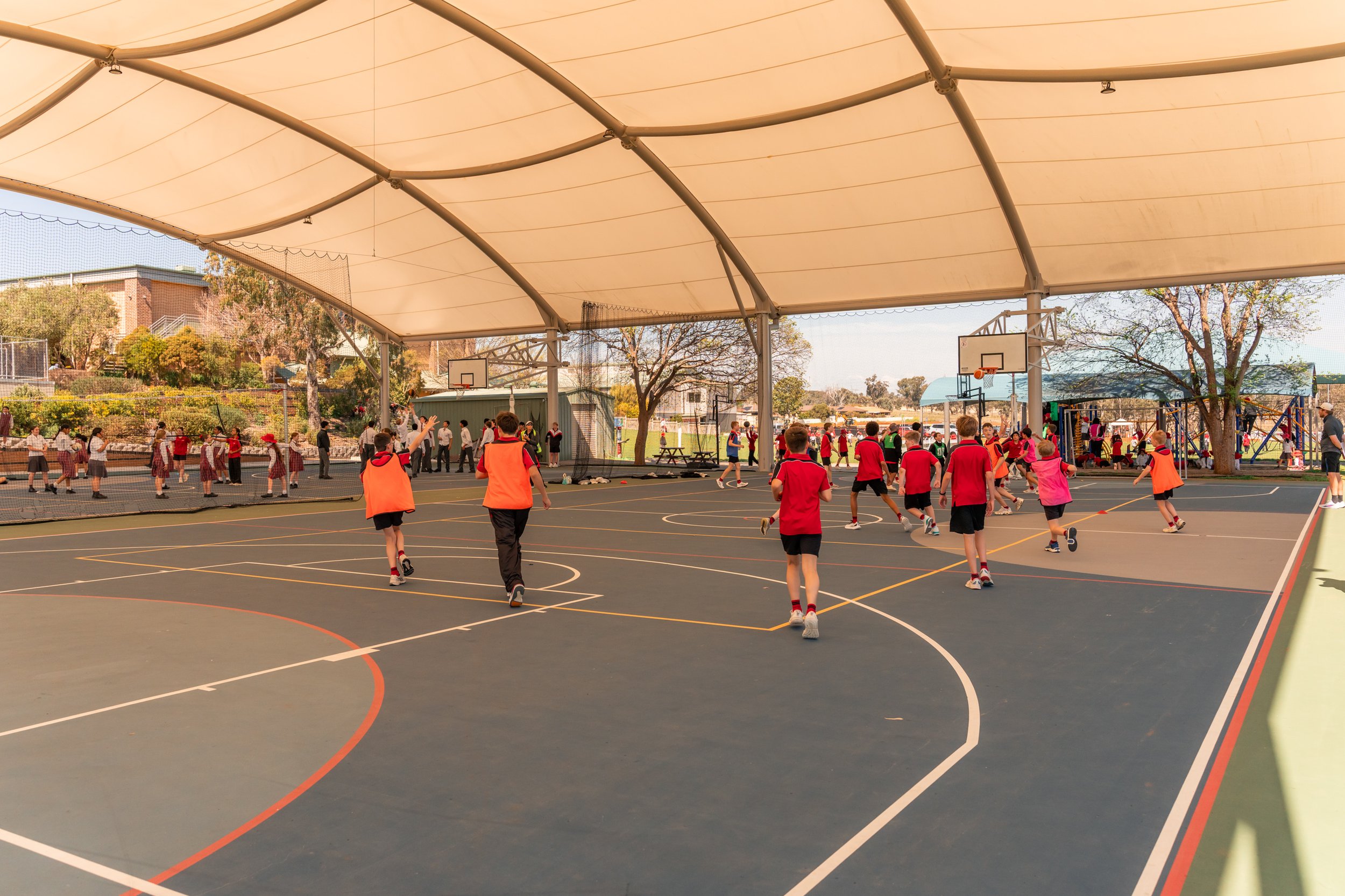 Children playing on an outdoor basketball court at a school under a large canopy during daytime.