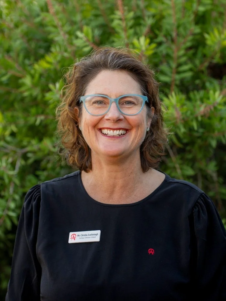 A woman with short brown hair, wearing glasses and a black shirt with a name badge, smiling in front of green foliage.