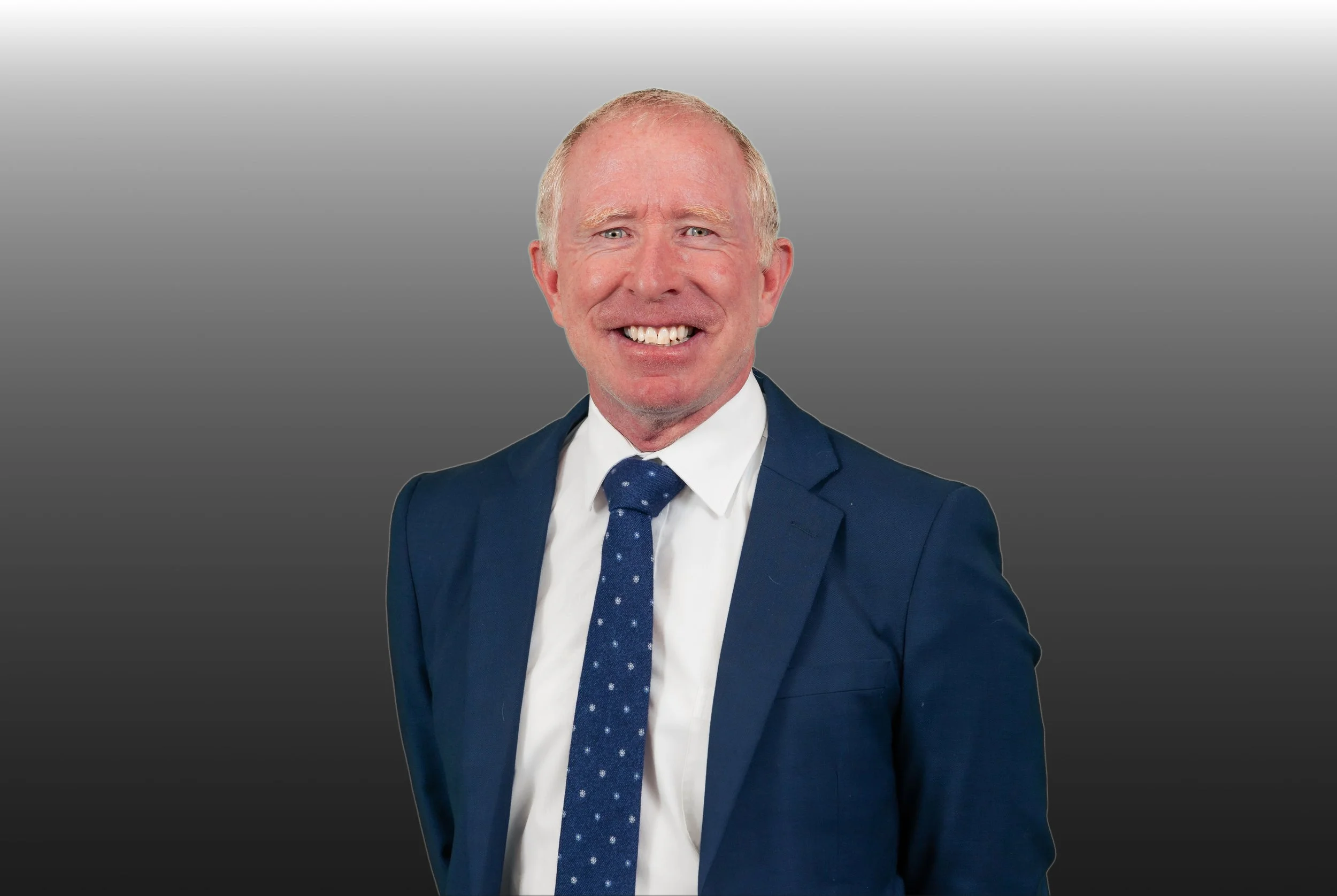 A smiling man in a navy blue suit, white shirt, and blue polka dot tie against a gray gradient background.