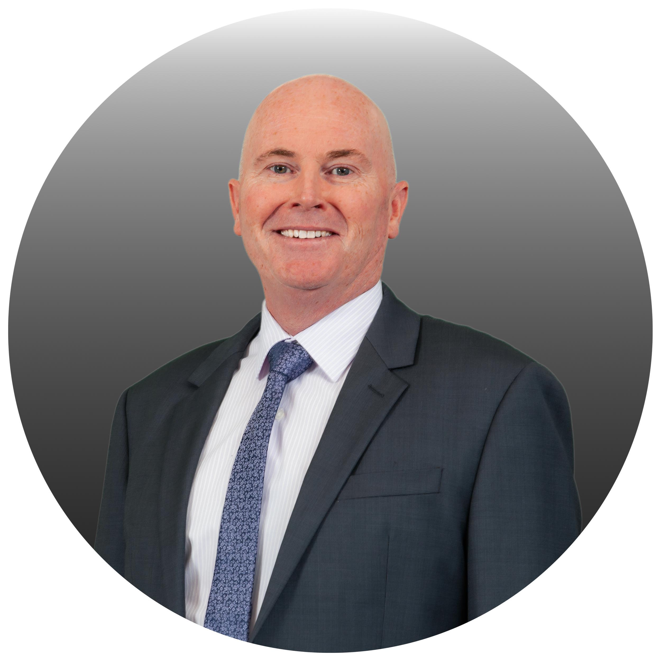 A professional man with a bald head and light skin, smiling, wearing a dark suit with a white shirt and a patterned blue tie, against a gradient grey background.