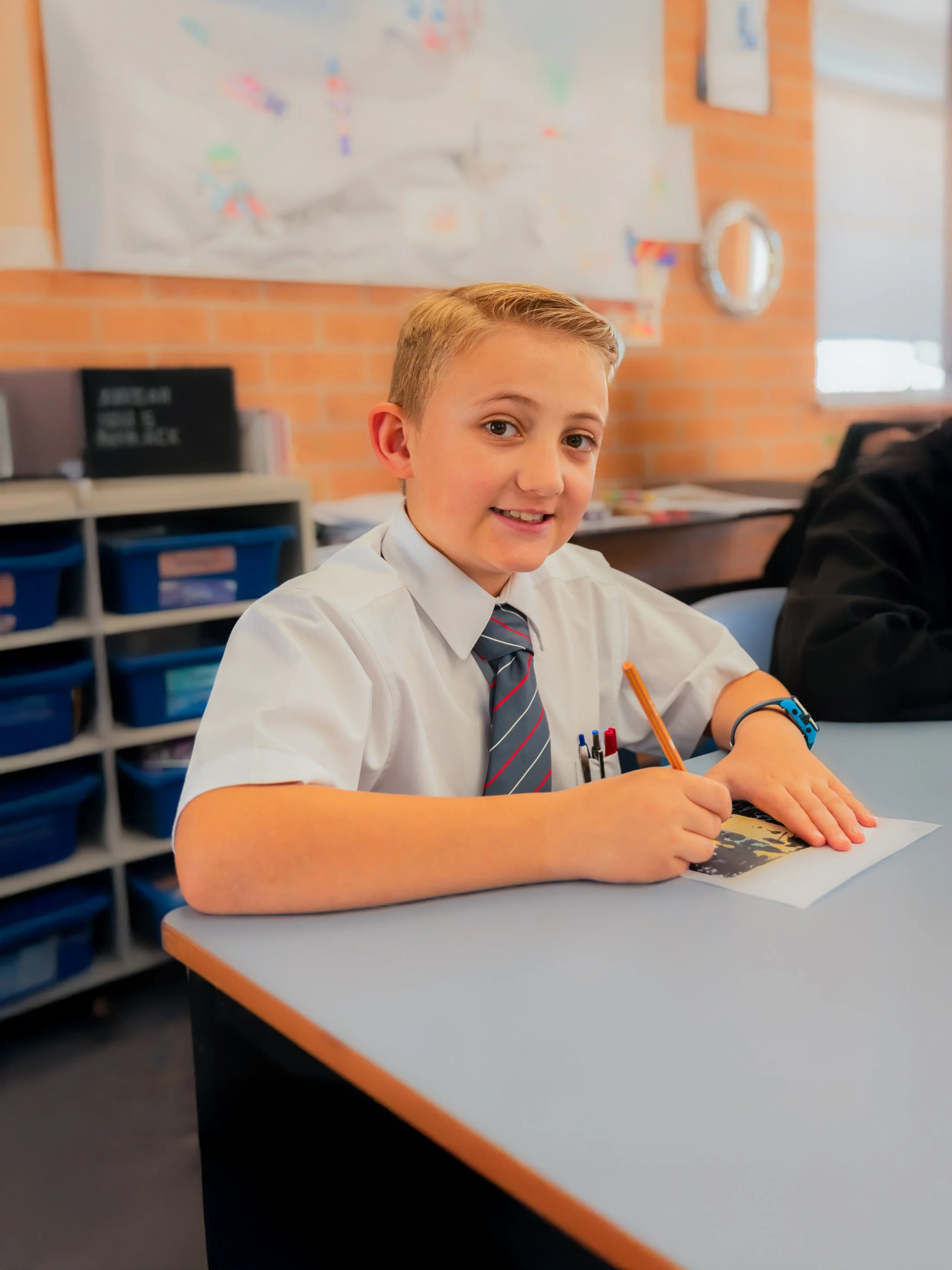 A young boy in school uniform smiling and writing at a desk in a classroom.