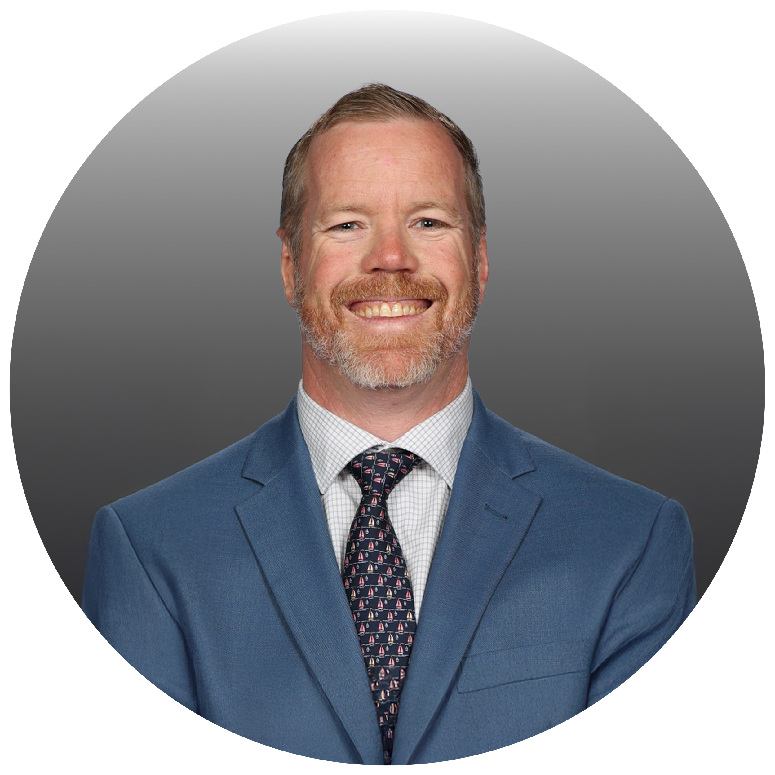 Portrait of a smiling man with a beard, wearing a blue suit, a checkered shirt, and a patterned tie, against a gray background.