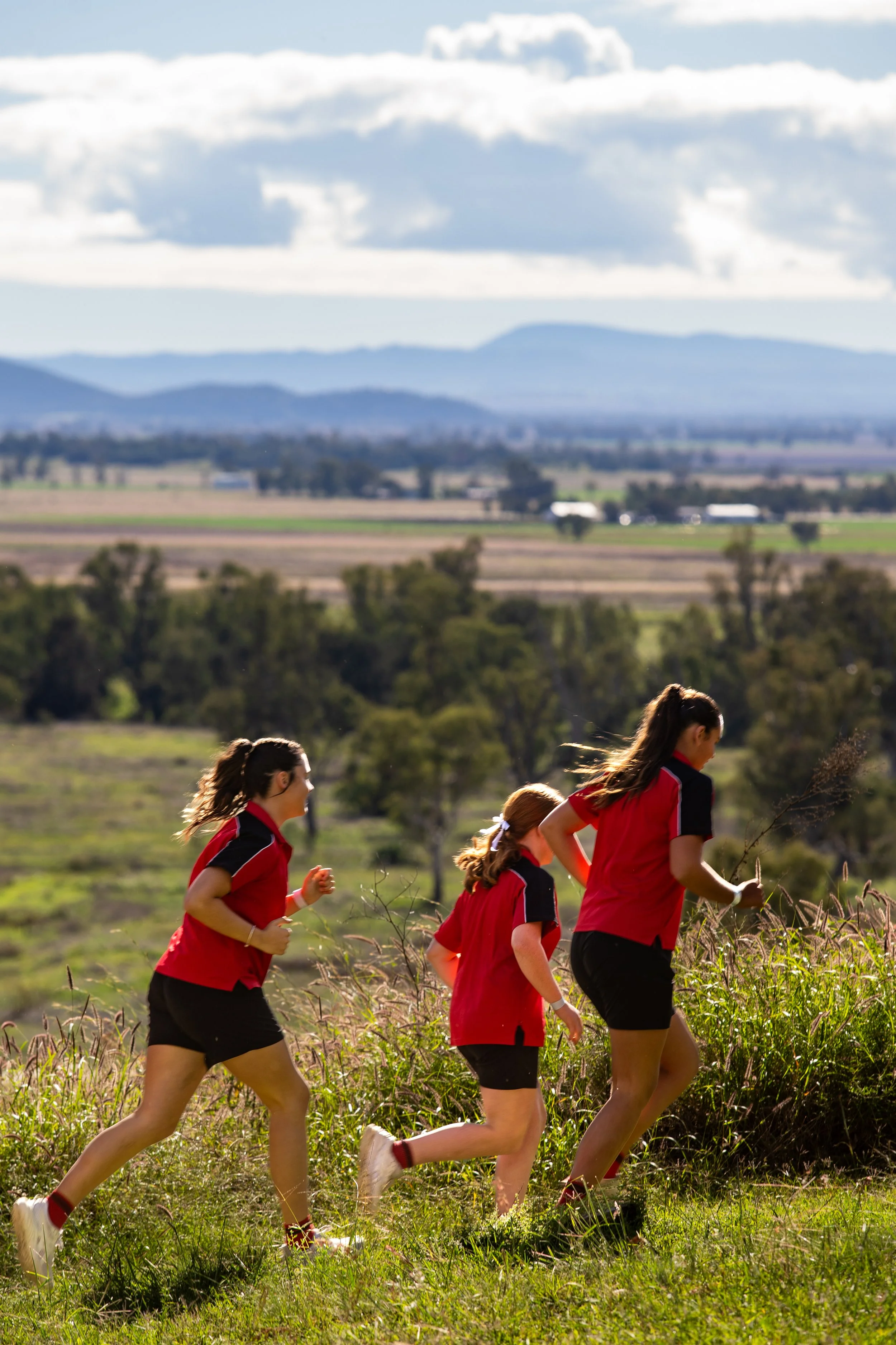 Four girls in red and black sports uniforms running outdoors on a grassy hillside with trees and distant mountains under a partly cloudy sky.