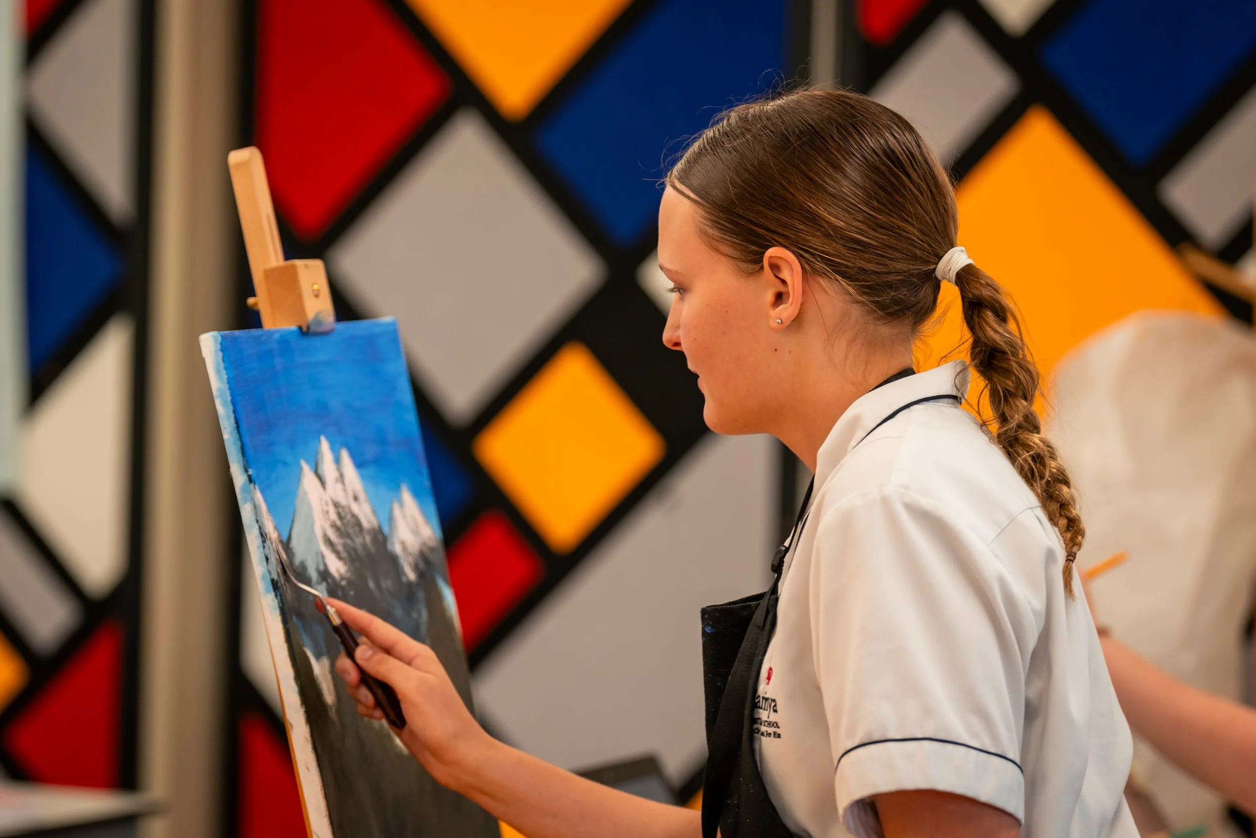 Carinya student with a braid wearing a white shirt and apron painting a mountain landscape on canvas, inside a Carinya Christian School, Gunnedah classroom.