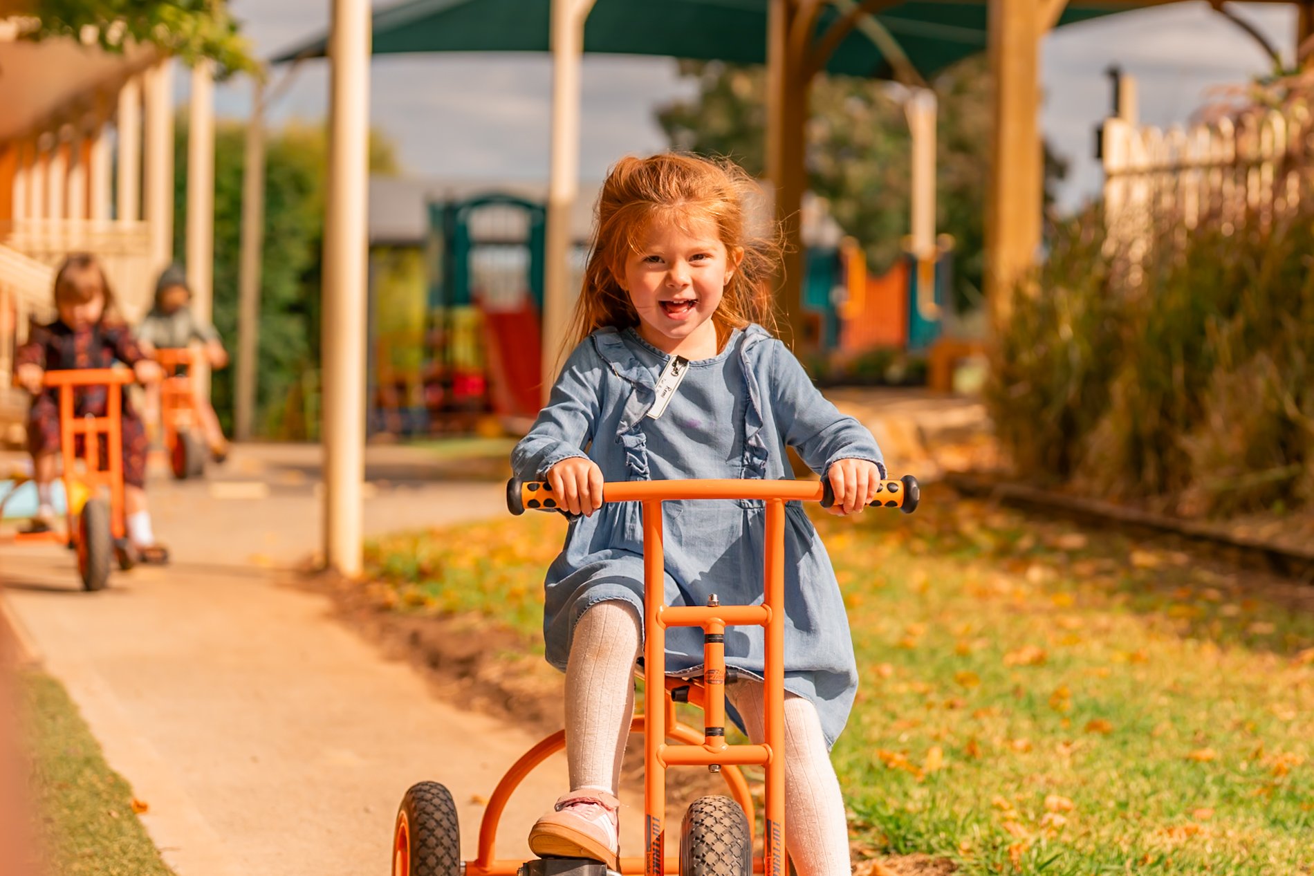 A Gumnuts student riding an orange tricycle outdoors at a playground, with other children and playground equipment in the background at Lilly Pilly Preps 3 Year Old Program in the outdoor play area at Carinya Christian School, Tamworth