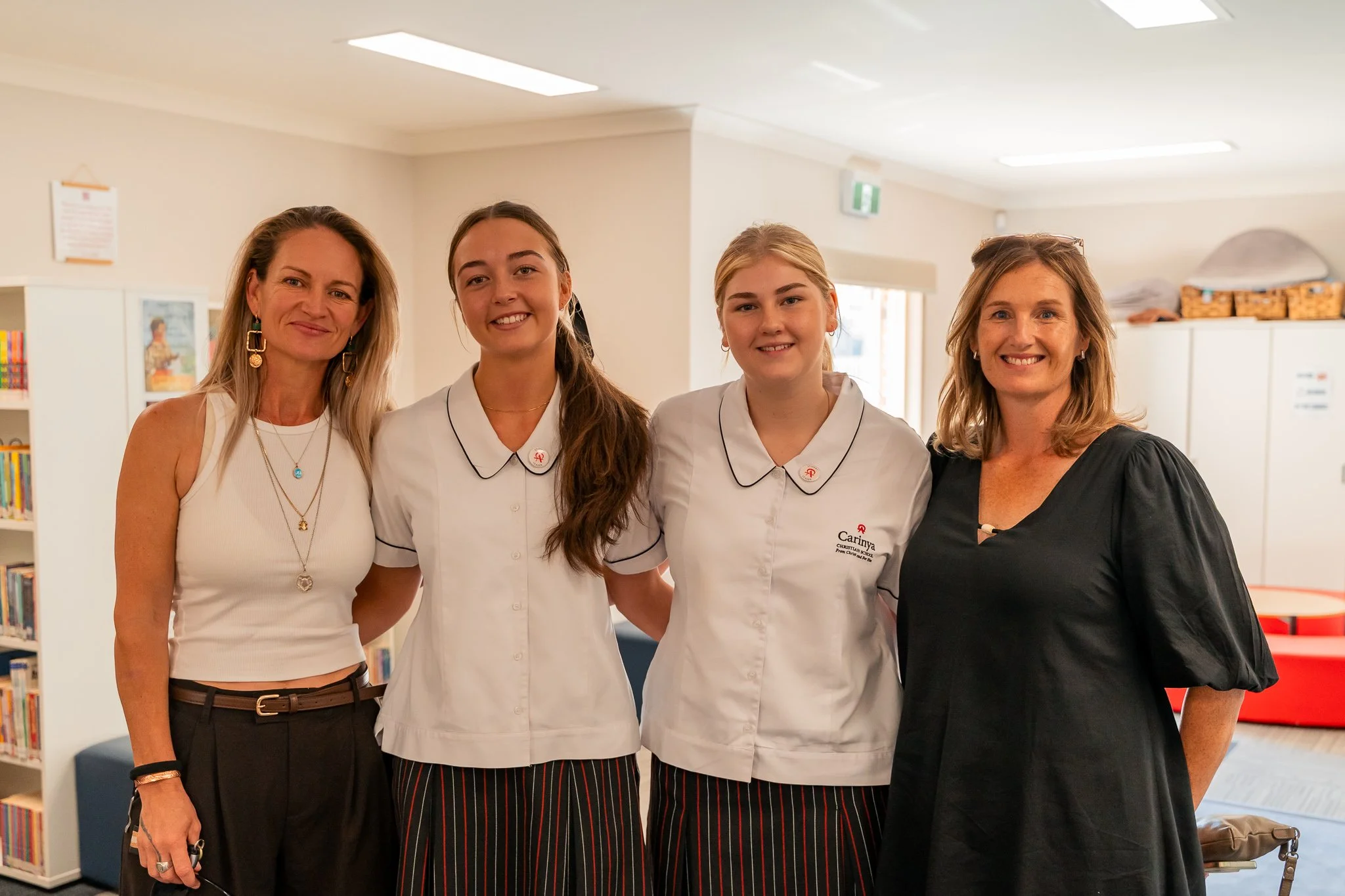Four women standing together indoors, two in school uniforms with white tops and striped skirts, and two in casual and black dresses, smiling at the camera.