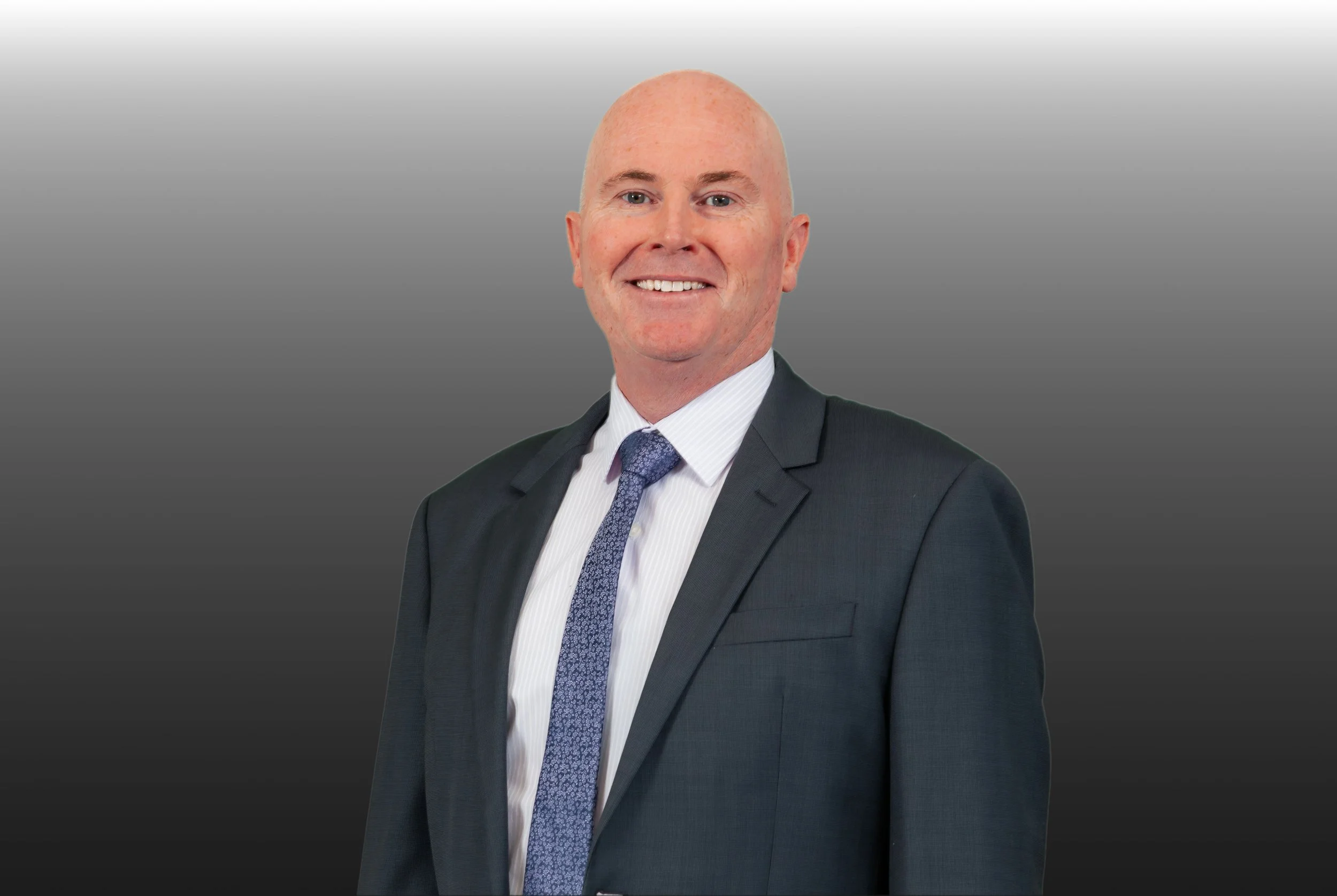 A man in a dark suit, white shirt, and patterned blue tie posing against a gray gradient background, smiling at the camera.