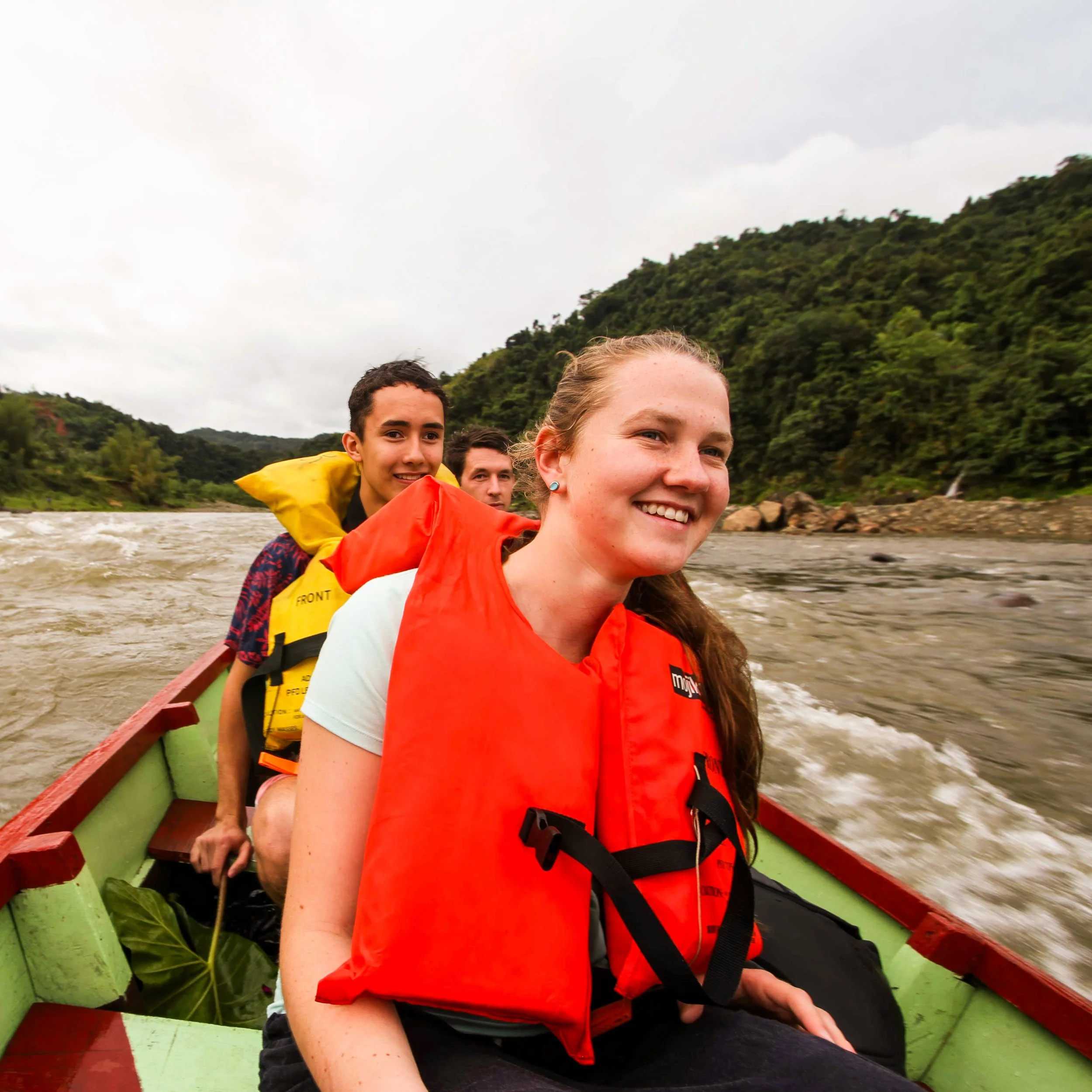 Three people wearing life jackets on a boat rowing on a river surrounded by green hills.