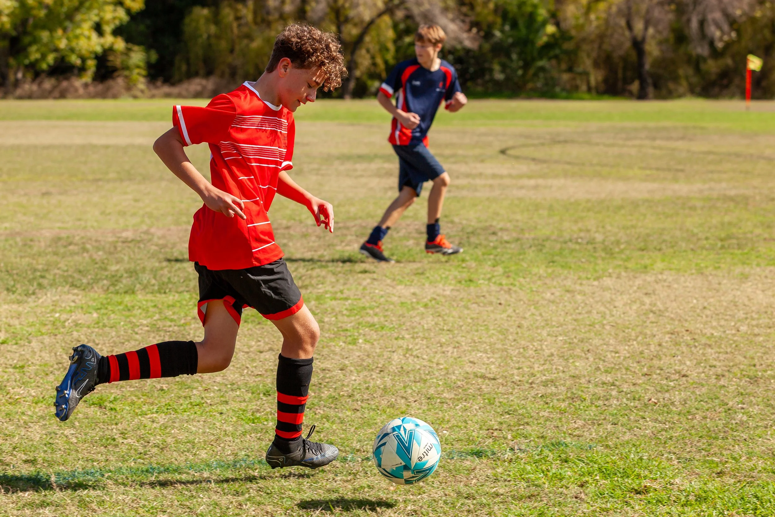 Two boys playing soccer on a grassy field on a sunny day, with trees in the background. The boy in the foreground is kicking a soccer ball, while the other is running behind him.