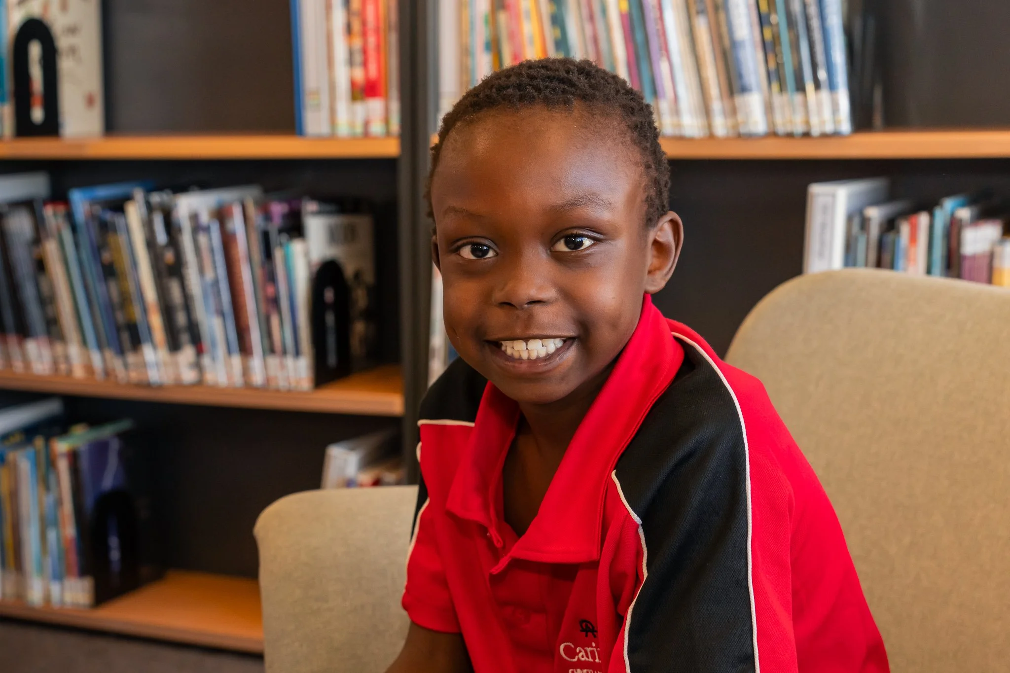 A young boy with short hair and a wide smile, wearing a red and black shirt, sitting in a library with shelves of books behind him.