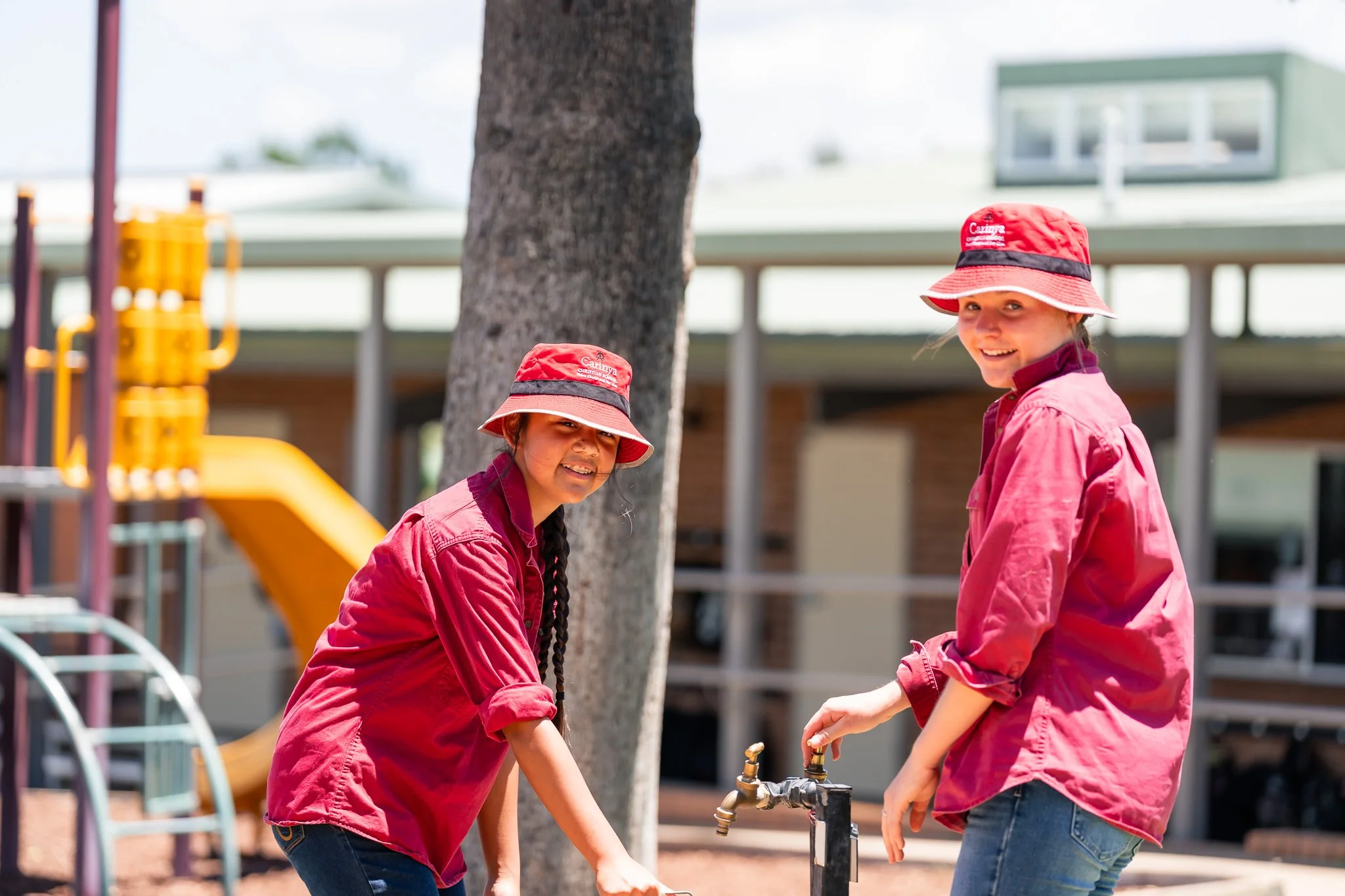 Two young girls wearing pink shirts and red hats turn on outdoor water faucets at a playground.