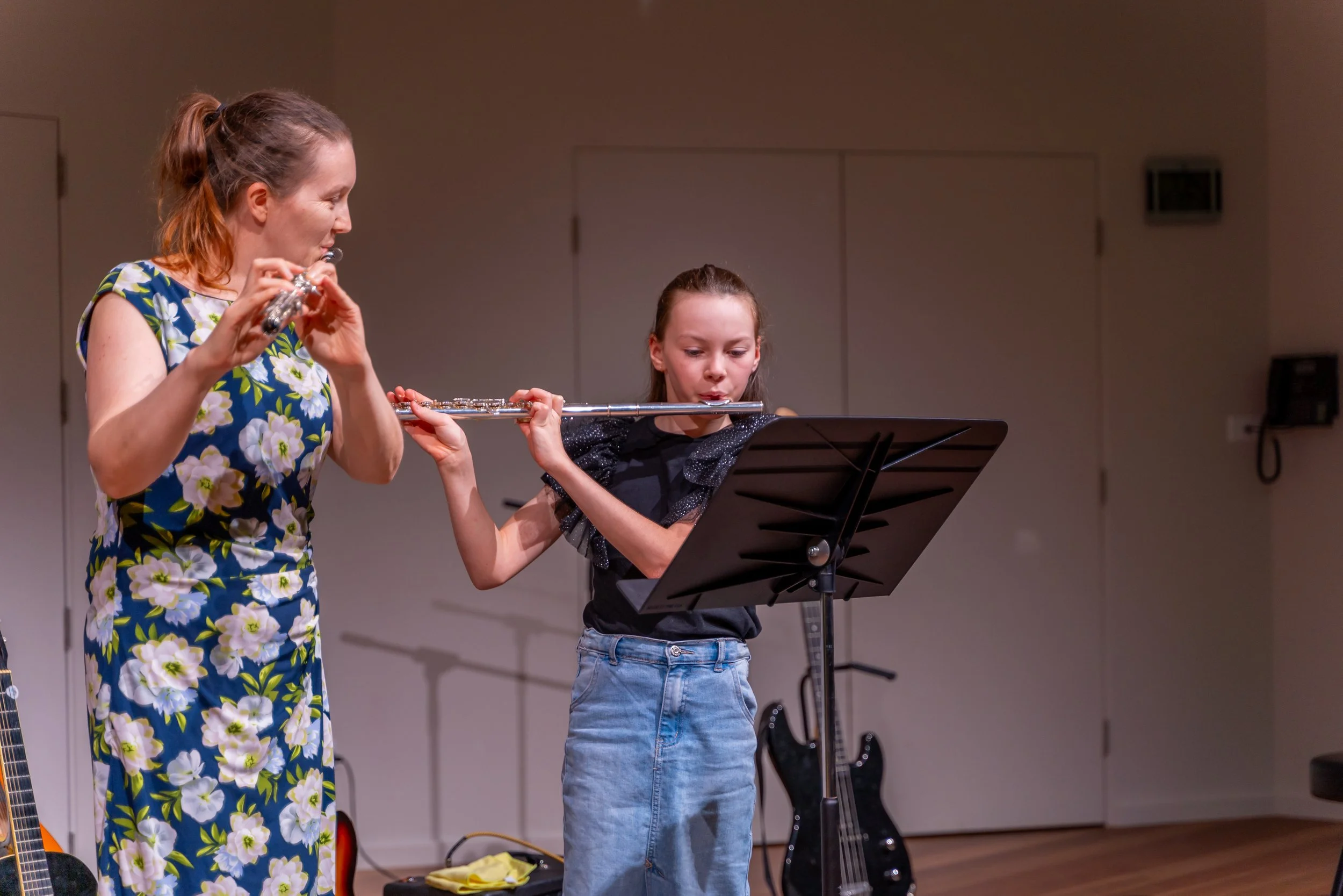 A woman teaching a young girl how to play the flute on stage, with music stands and guitars in the background.