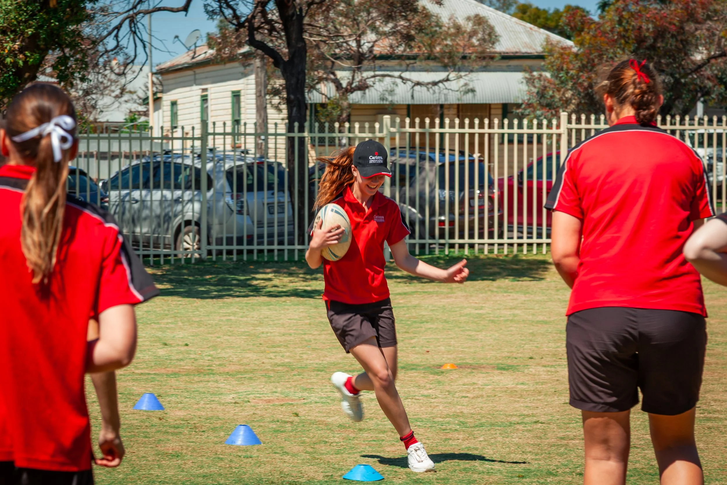 Young woman running on the grass field with a rugby ball, participating in a sports activity or training session with other women around, on a sunny day.