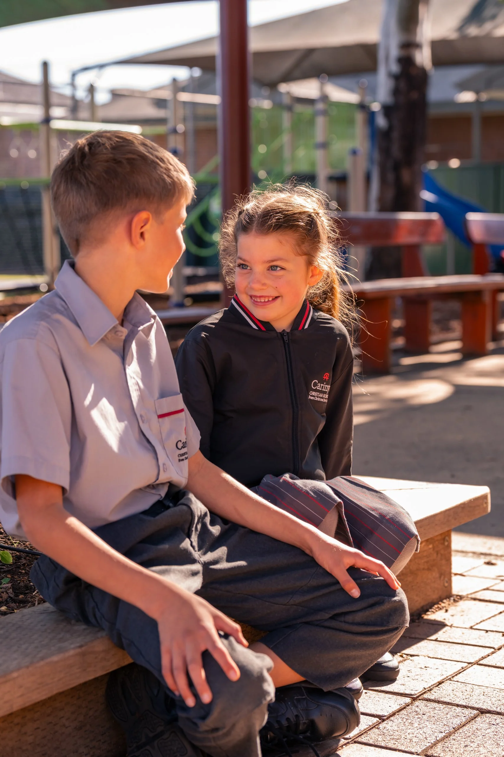 A boy and girl sitting on a park bench engaging in conversation, smiling, with a playground in the background.