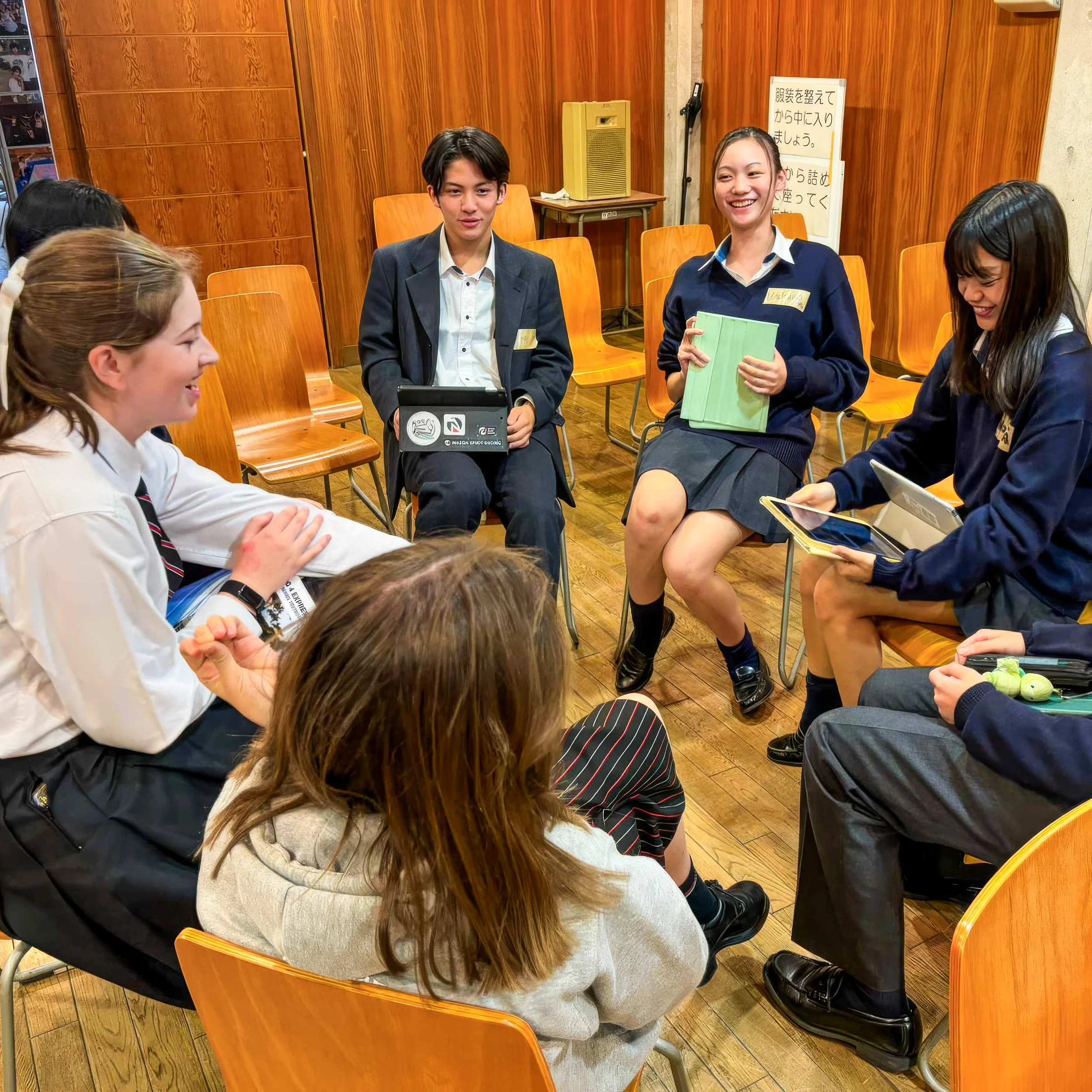 Group of school students sitting in a circle, laughing, talking, and using tablets and notebooks during a class or discussion.