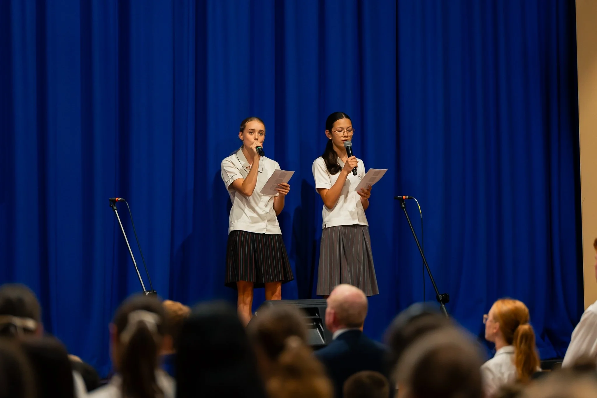 Two young women stand on stage in front of a blue curtain, holding papers and speaking into microphones during a presentation or performance. Audience members are visible in the foreground.