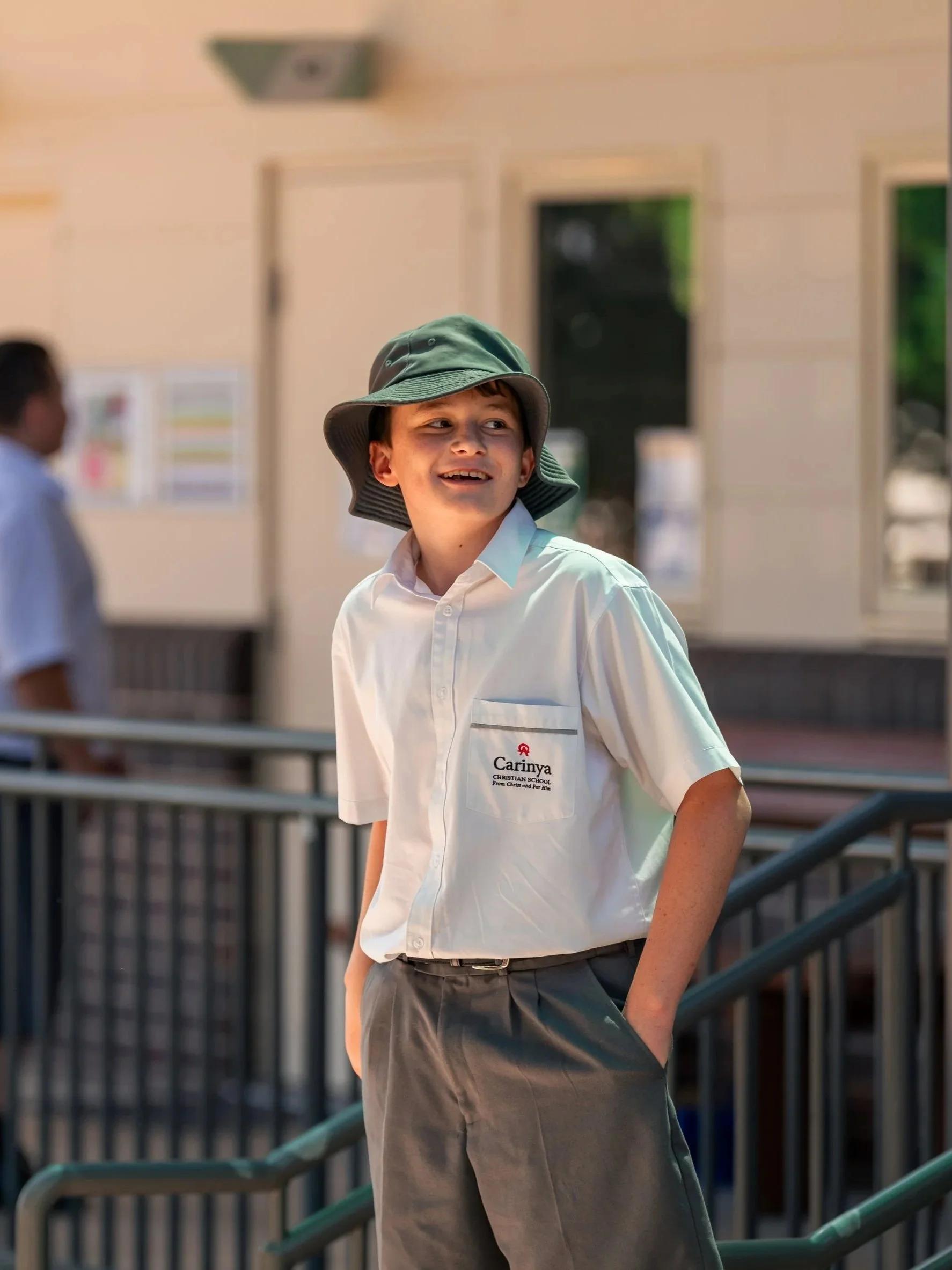 A young boy in a school uniform with hands in pockets, wearing a green bucket hat and smiling, standing outside a school building with other students in the background.