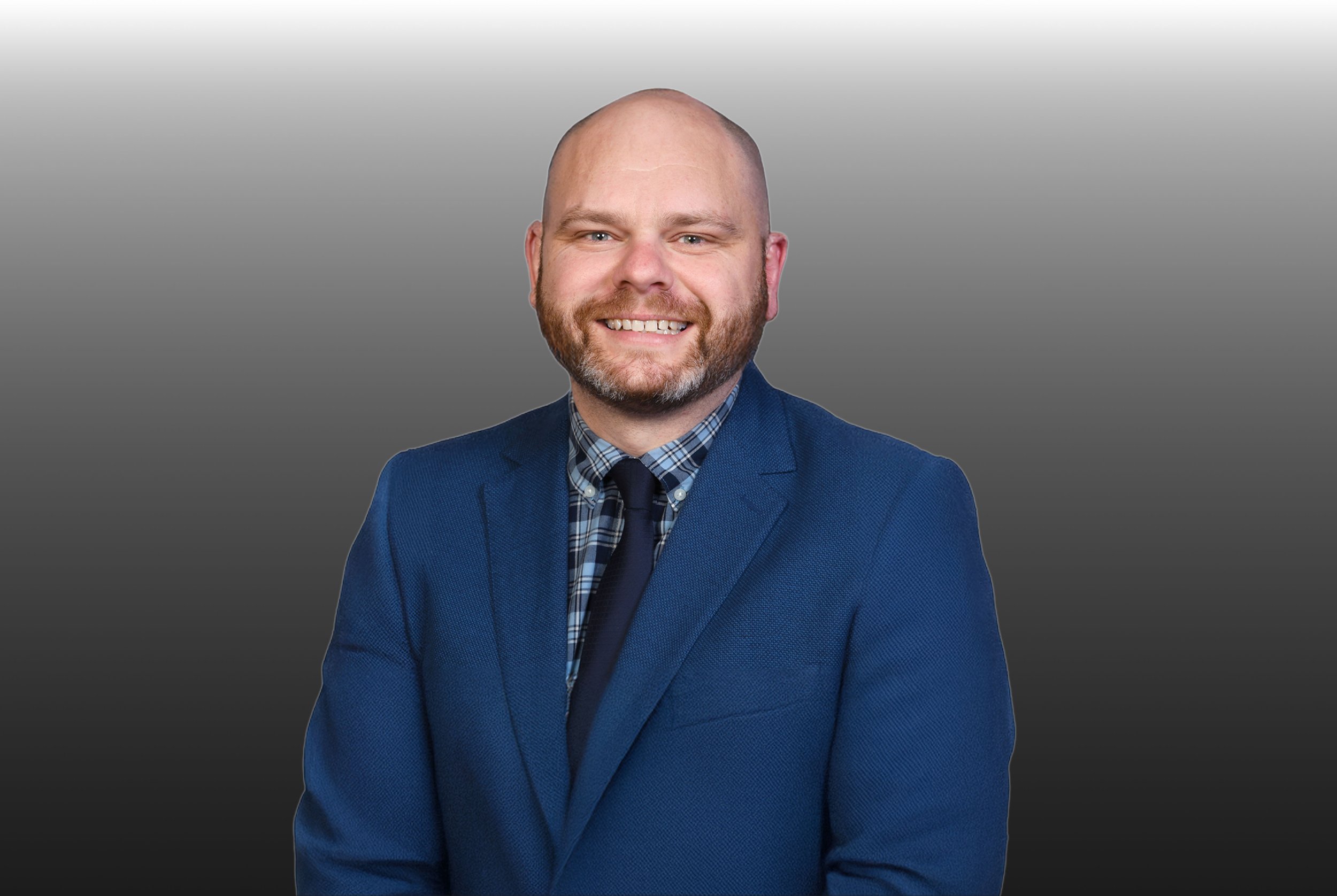 A man with a beard in a blue suit blazer, plaid shirt, and dark tie, smiling against a gradient background.