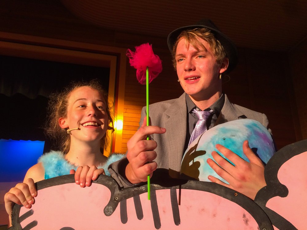 Two young actors on stage, one girl smiling and one boy holding a flower and a painted ball, performing in a play with colorful props and costumes.