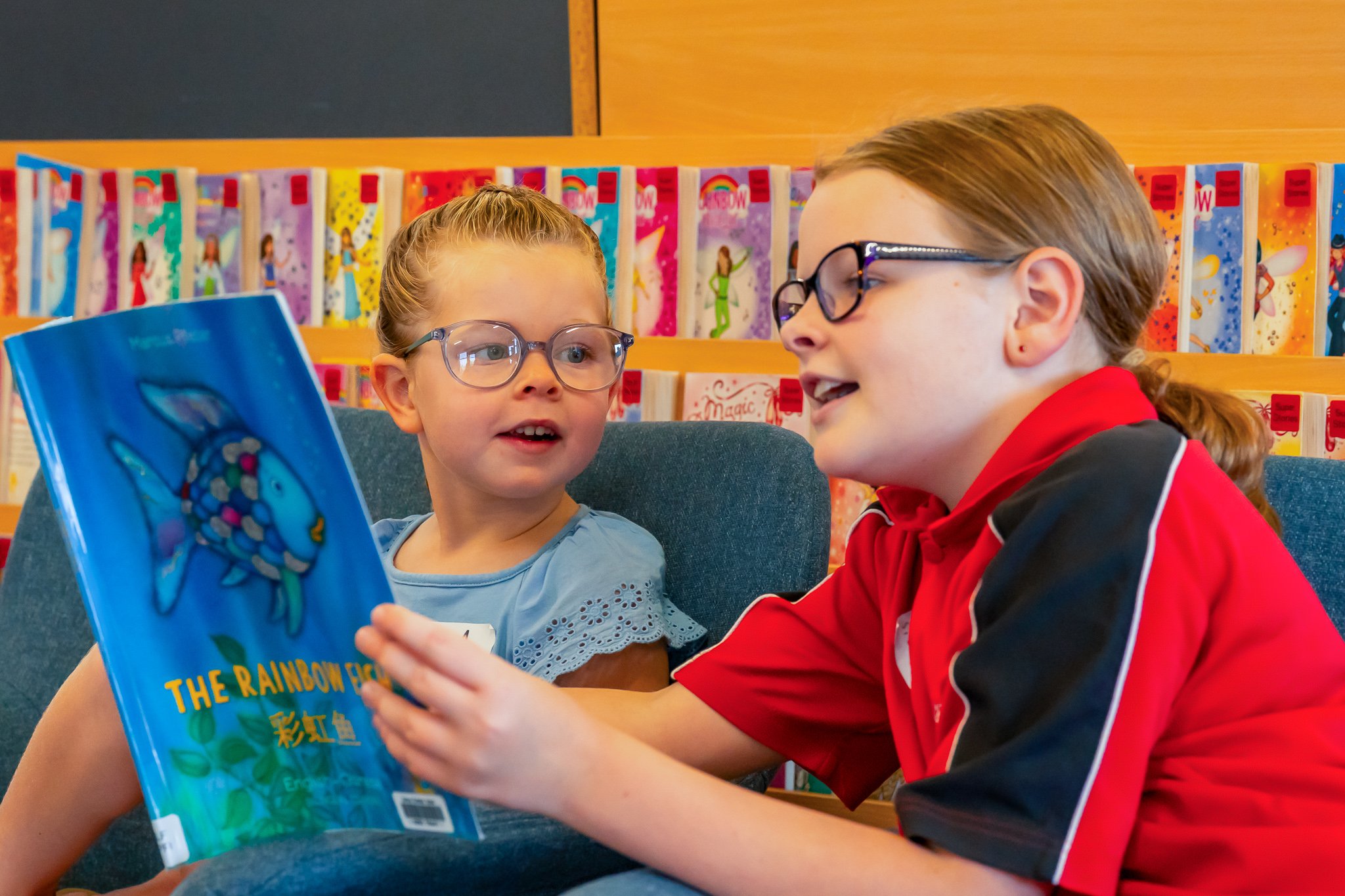 Two young girls wearing glasses sitting on a blue couch in a bookstore or library, looking at each other, with one girl holding a colorful book titled 'The Rainbow Fish'.