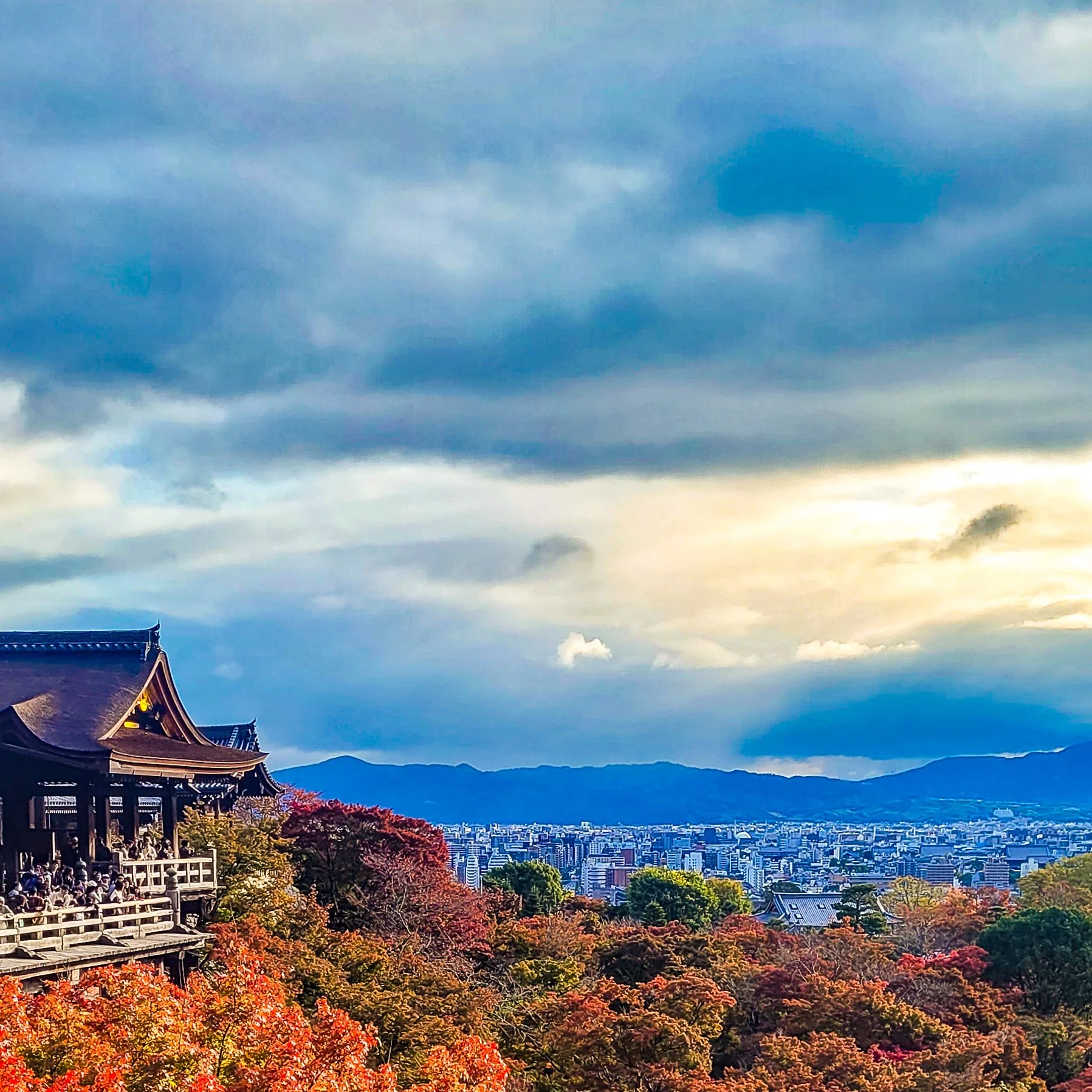 Scenic view of a Japanese temple on a hillside with autumn trees, overlooking a city with mountains in the background under a cloudy sky.