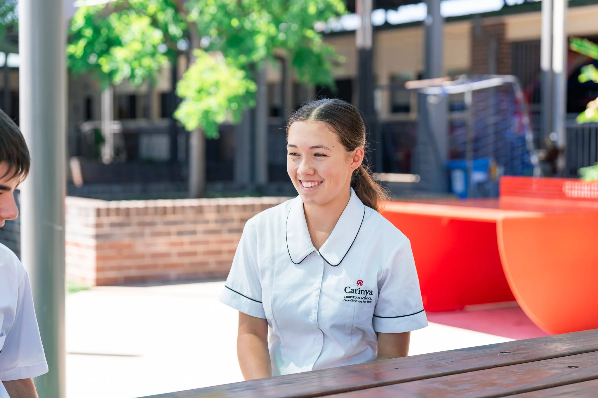 A teenage girl smiling while sitting outdoors at a wooden picnic table, wearing a white school uniform with a crest on the pocket that says 'Carinya Christian School'.