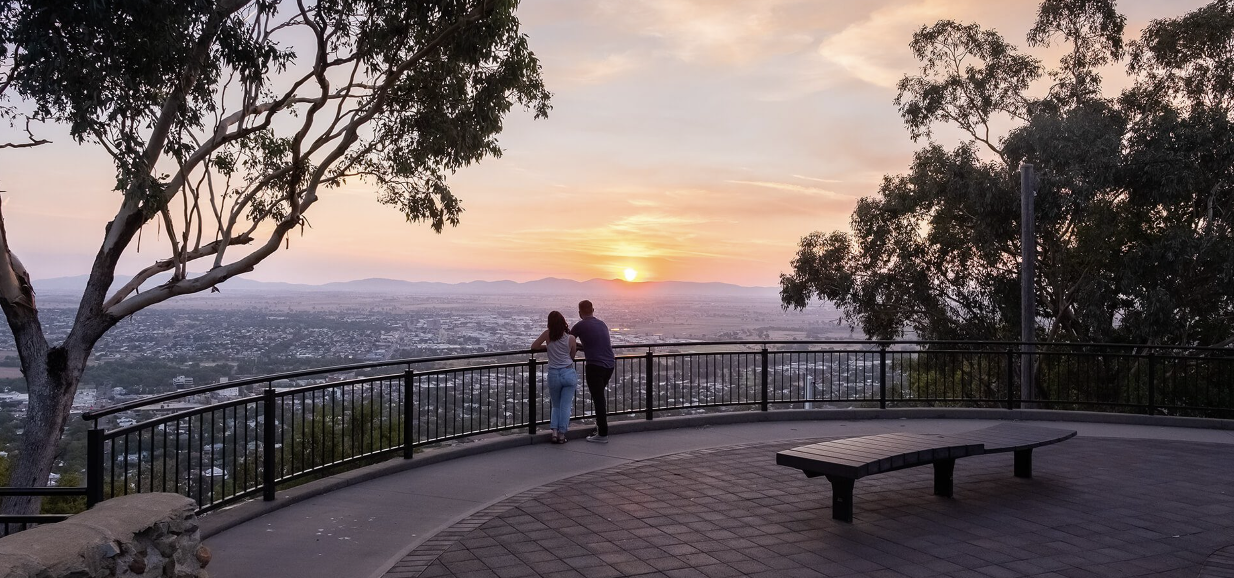 A couple standing on an observation deck, watching a sunset over a city with distant mountains, surrounded by trees and a metal railing.