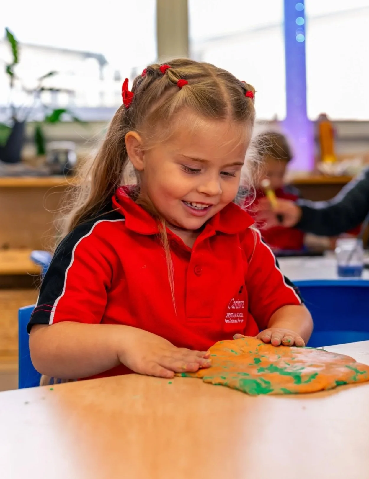 A young girl in a red school uniform playing with colorful slime on a table in a classroom.