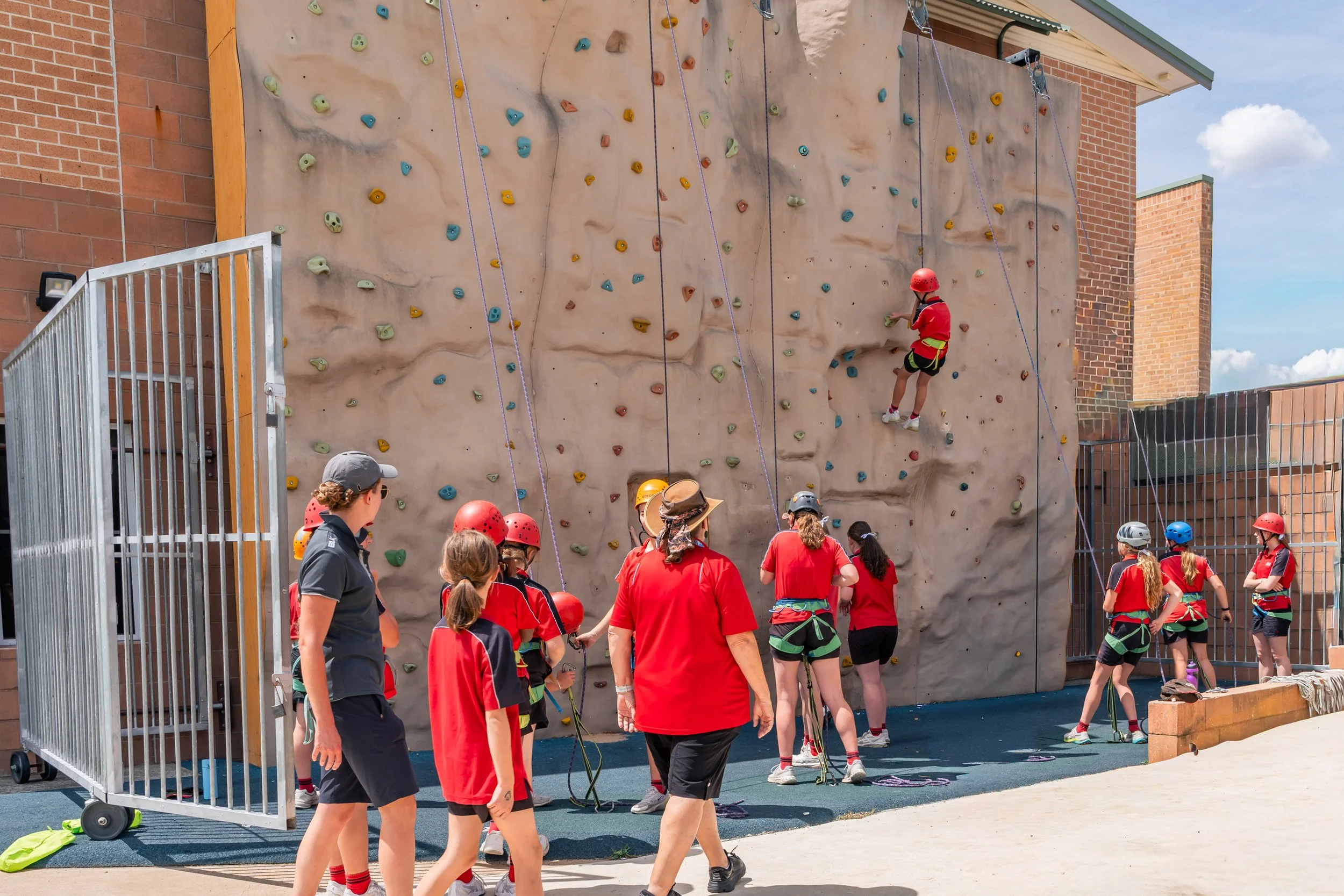 A group of children and adults participating in a rock climbing activity outdoors, with some children climbing on a rock wall and others waiting their turn, all wearing helmets and harnesses.