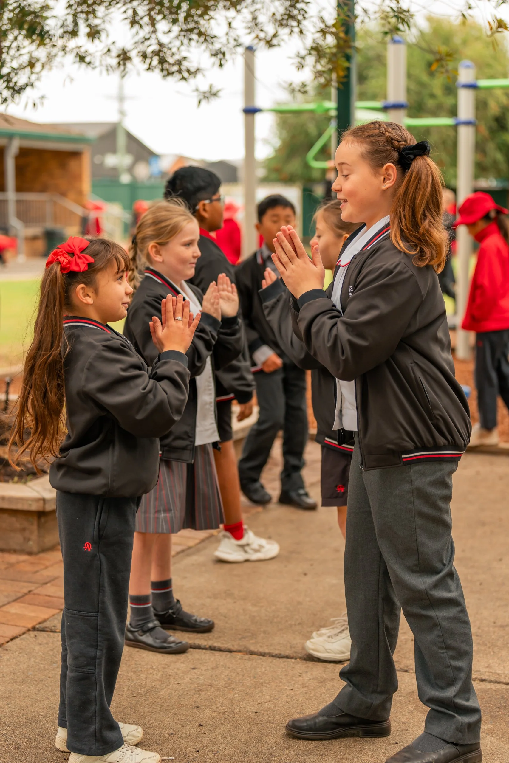 Group of children in school uniforms, some with red bows or caps, practicing a greeting with hands pressed together outdoors on school grounds.