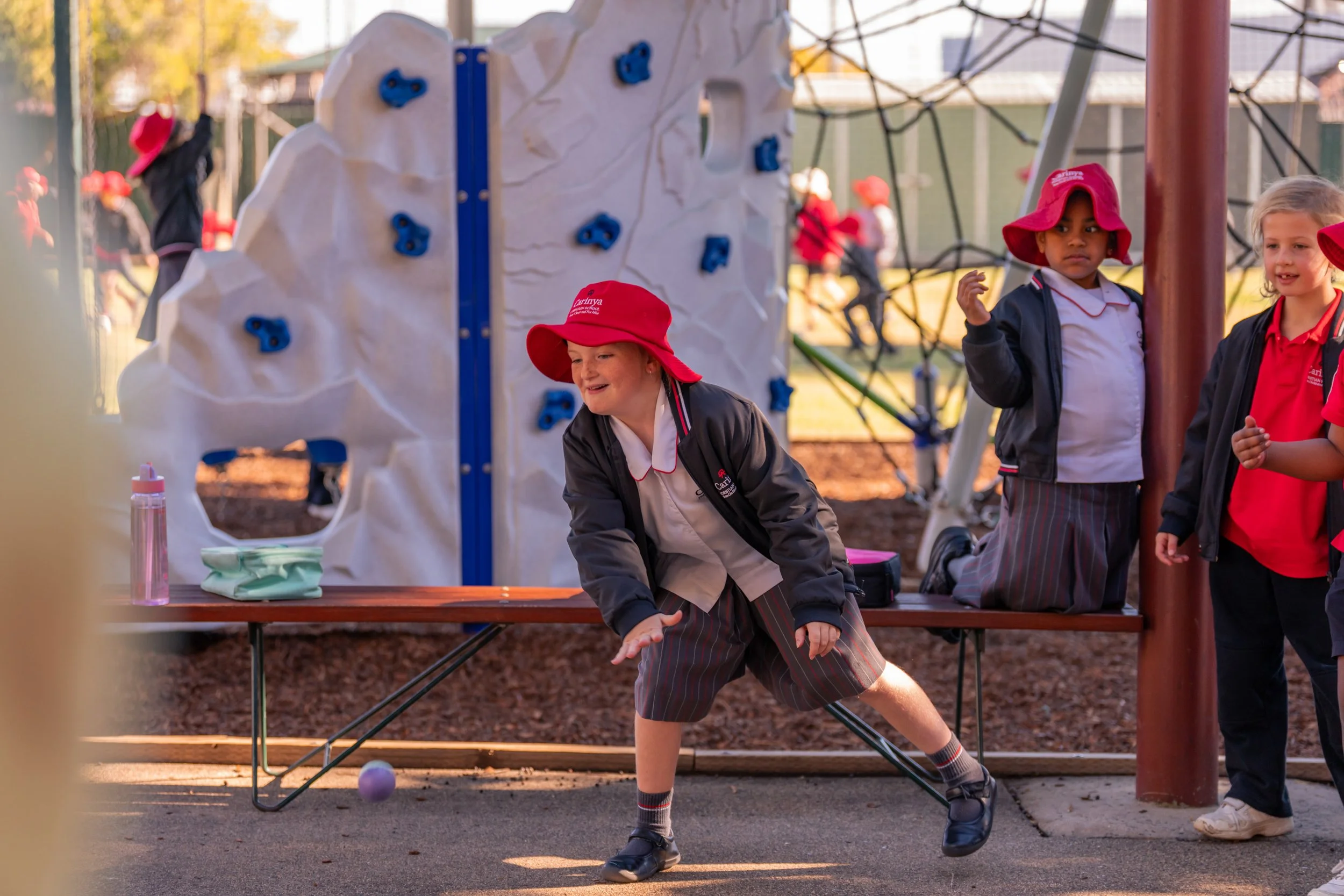 Children in school uniforms playing outside on a playground, one child is throwing a ball while others watch, with playground equipment in the background.