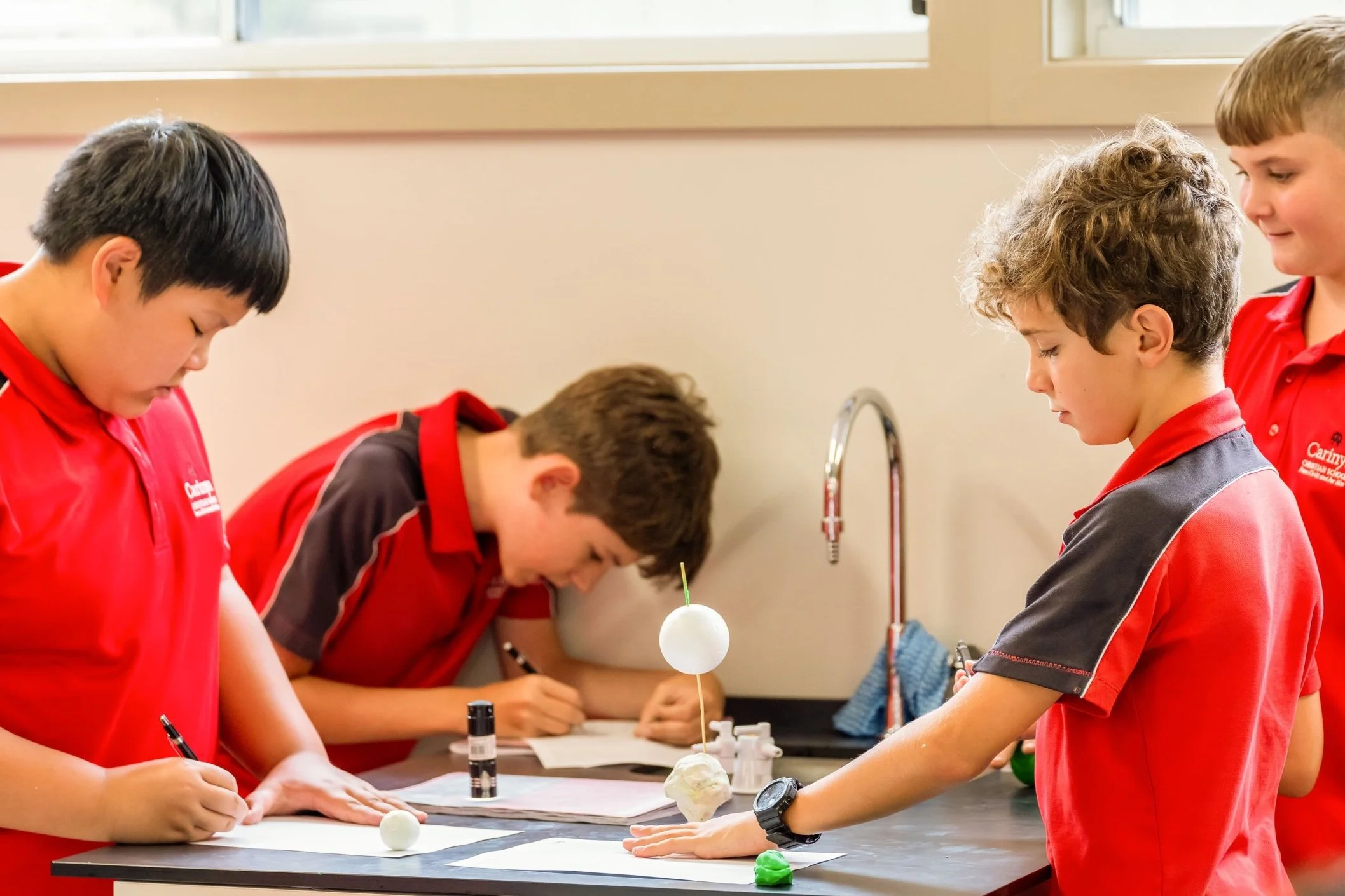 Four children in red and gray uniforms working on a science experiment at a laboratory table.