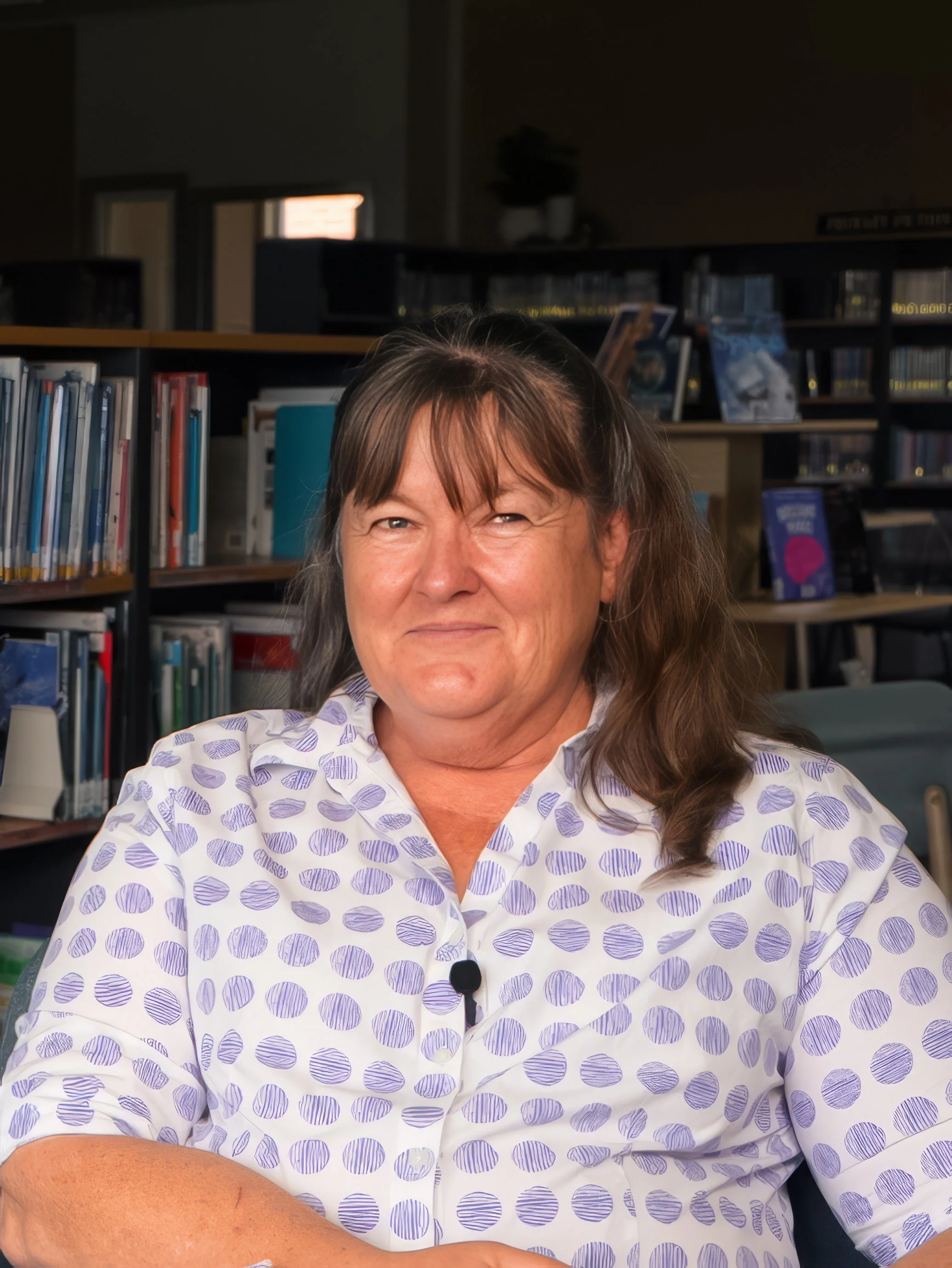 A woman with brown hair wearing a white shirt with purple polka dots sitting in a library.
