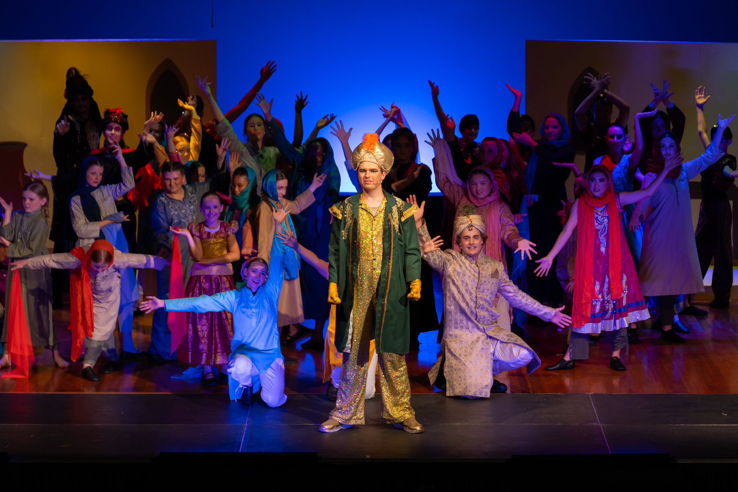 A group of children in colorful costumes on stage, performing a play or dance. The front center features a child dressed as a prince or sultan with a turban and gold outfit. The background shows a set resembling a Middle Eastern or Arabian setting.