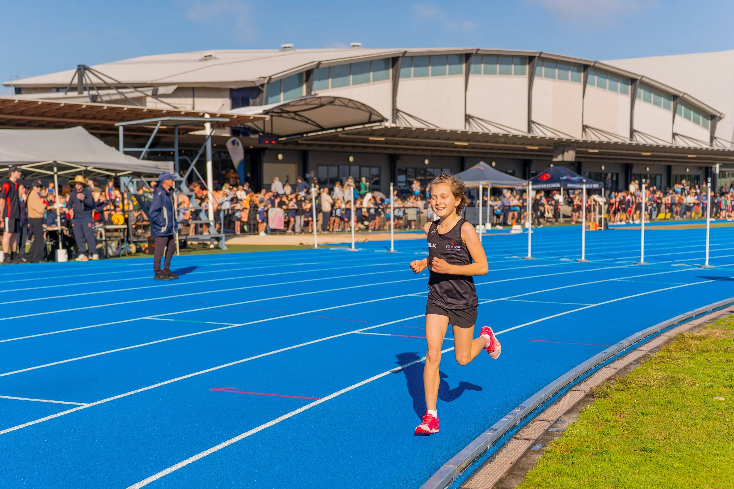 A young girl running on a blue track at an outdoor stadium during an athletic event, with a large crowd of spectators and officials in the background.