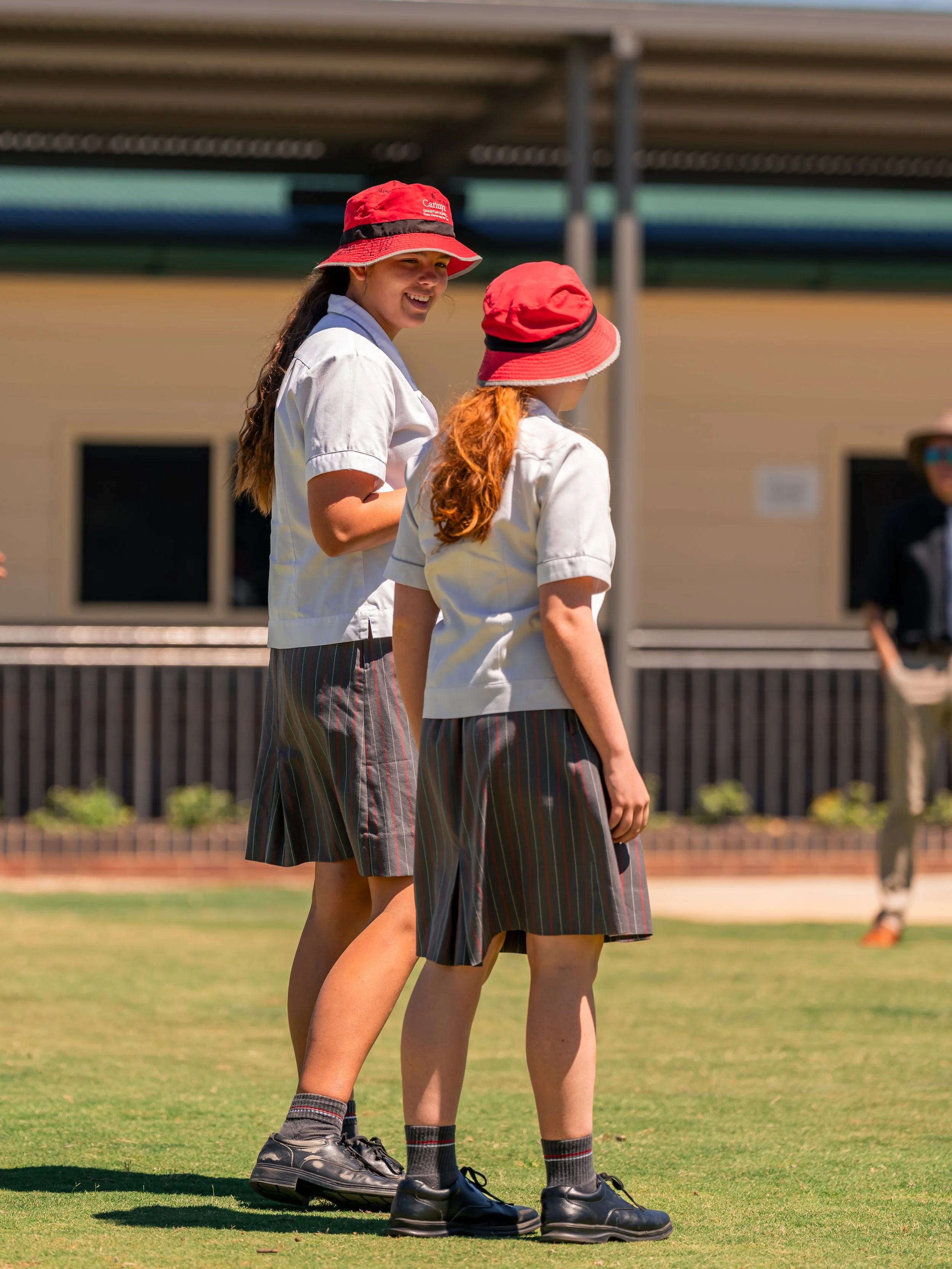 Two Carinya students in uniform standing and talking outdoors on a large sports oval, wearing red hats, white shirts, and striped skirts with black shoes at Carinya Christian School, Gunnedah.
