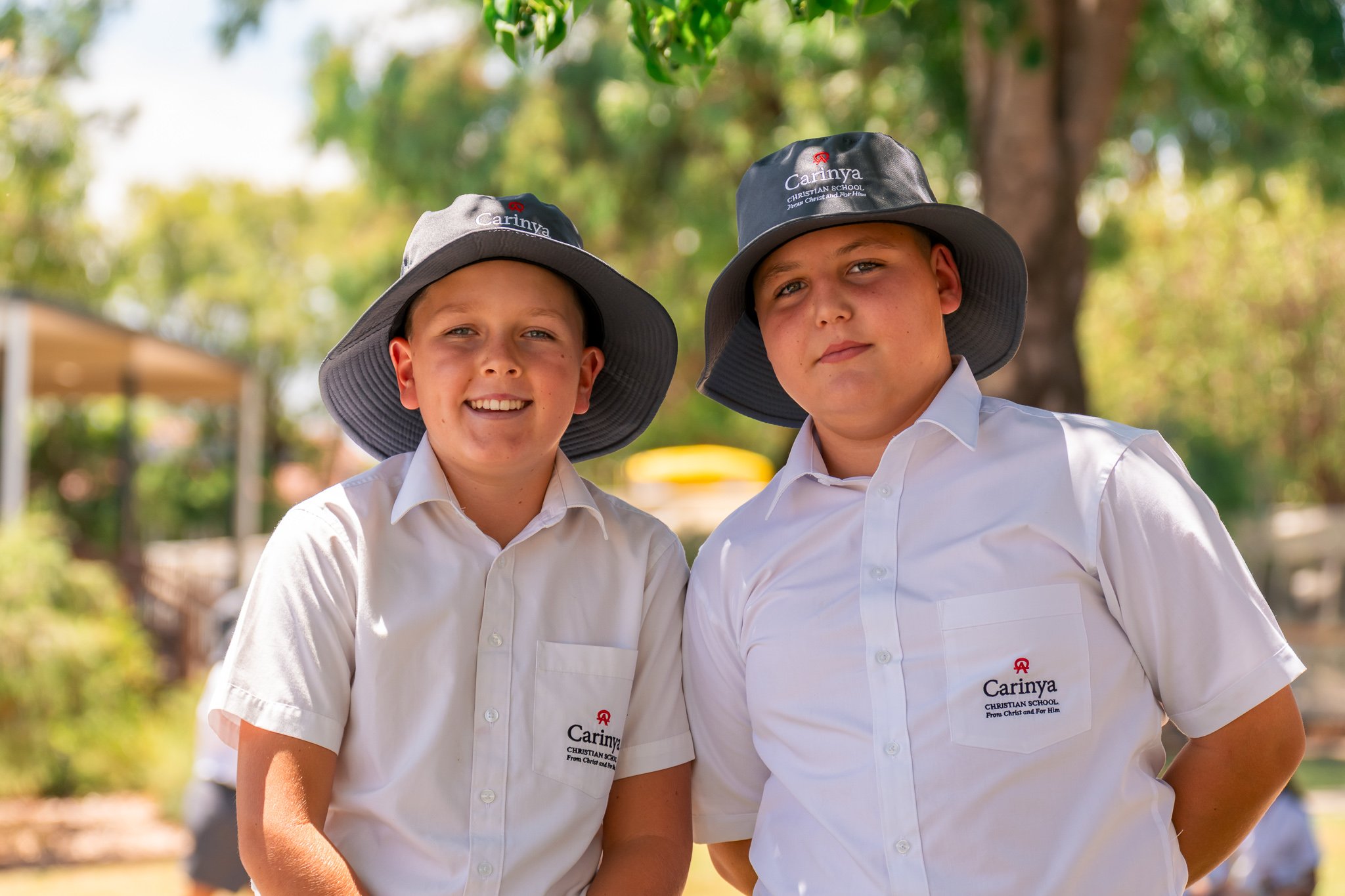 Two young boys wearing white school shirts and wide-brimmed gray hats with the word 'Carinya' on them, standing outdoors with trees and a yellow object in the background.