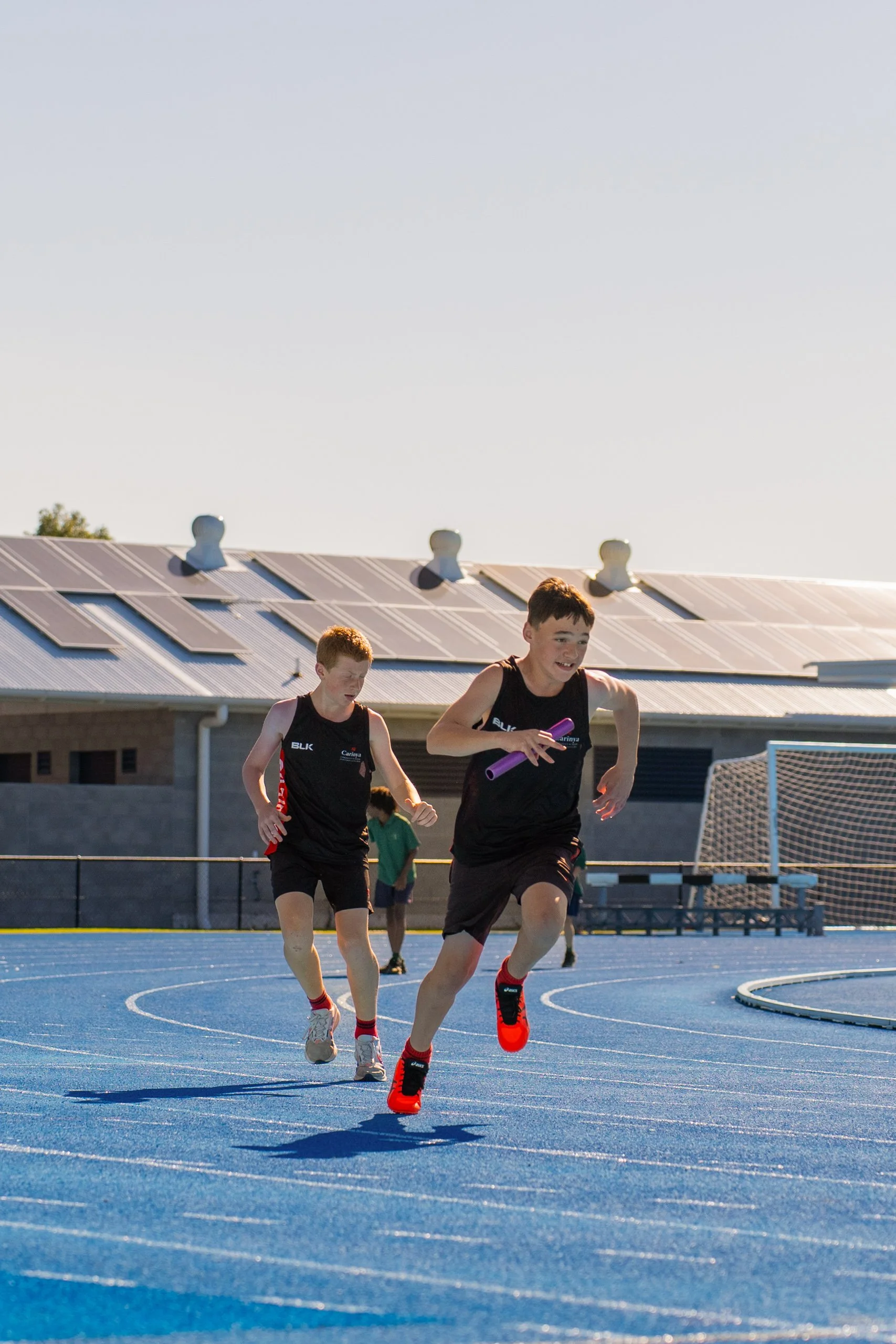 Two boys running on a blue track during daytime, with a building and solar panels in the background.