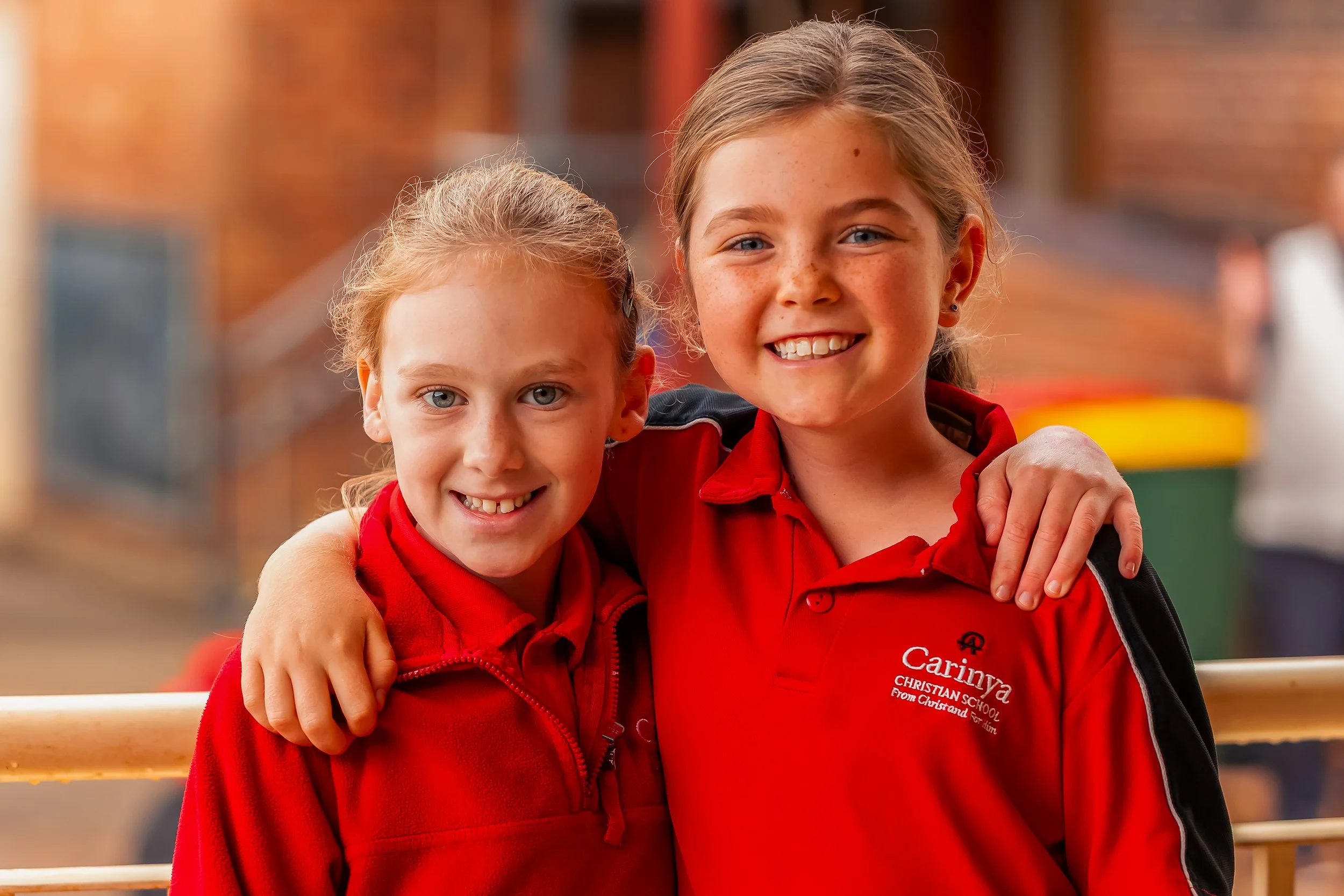 Two young girls wearing red school uniforms smiling and standing close with their arms around each other, outdoors at a school.