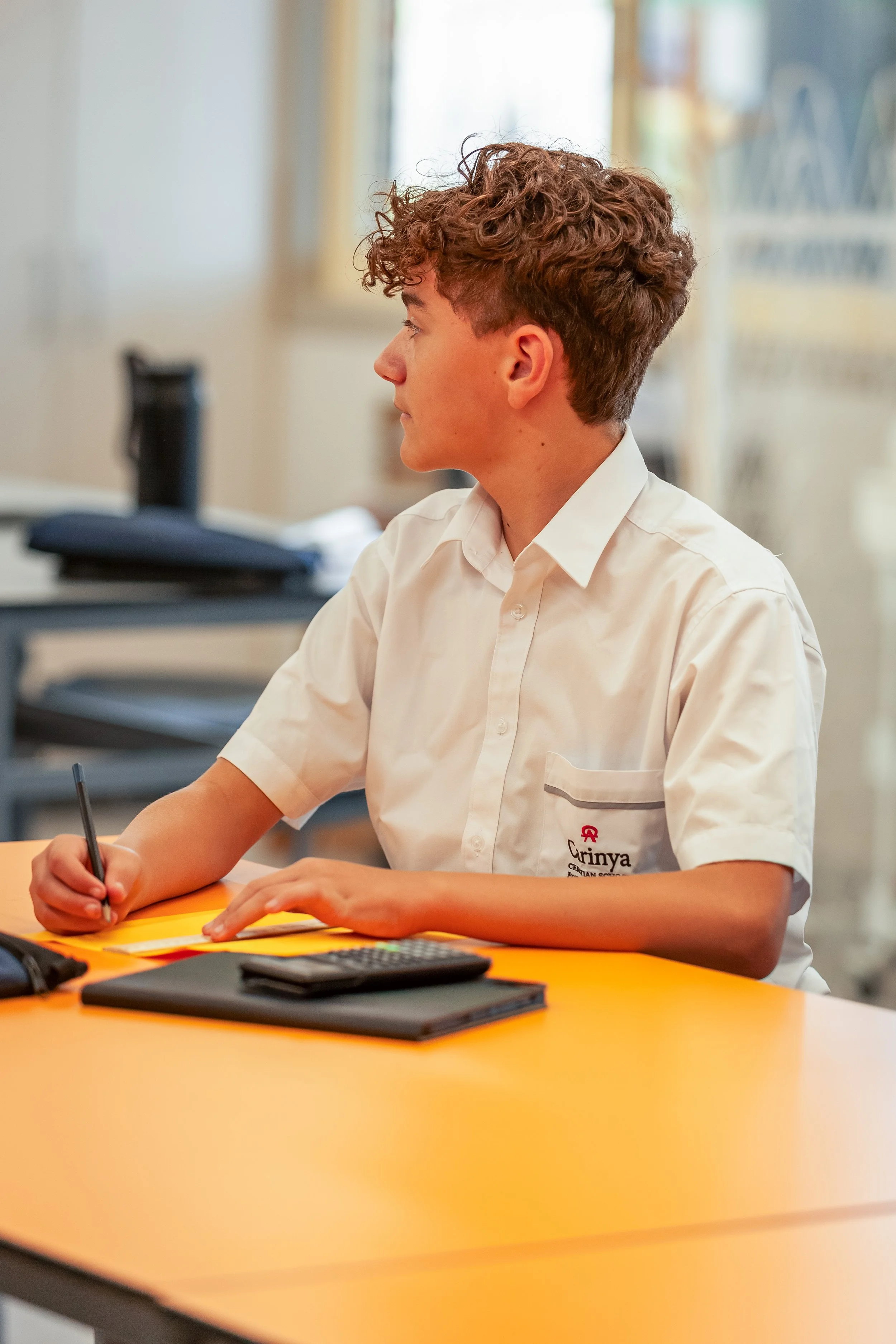A young male student with curly hair, wearing a white school uniform shirt, sitting at a yellow table in a classroom, writing in a notebook with a pen, with a calculator and stack of papers in front of him.