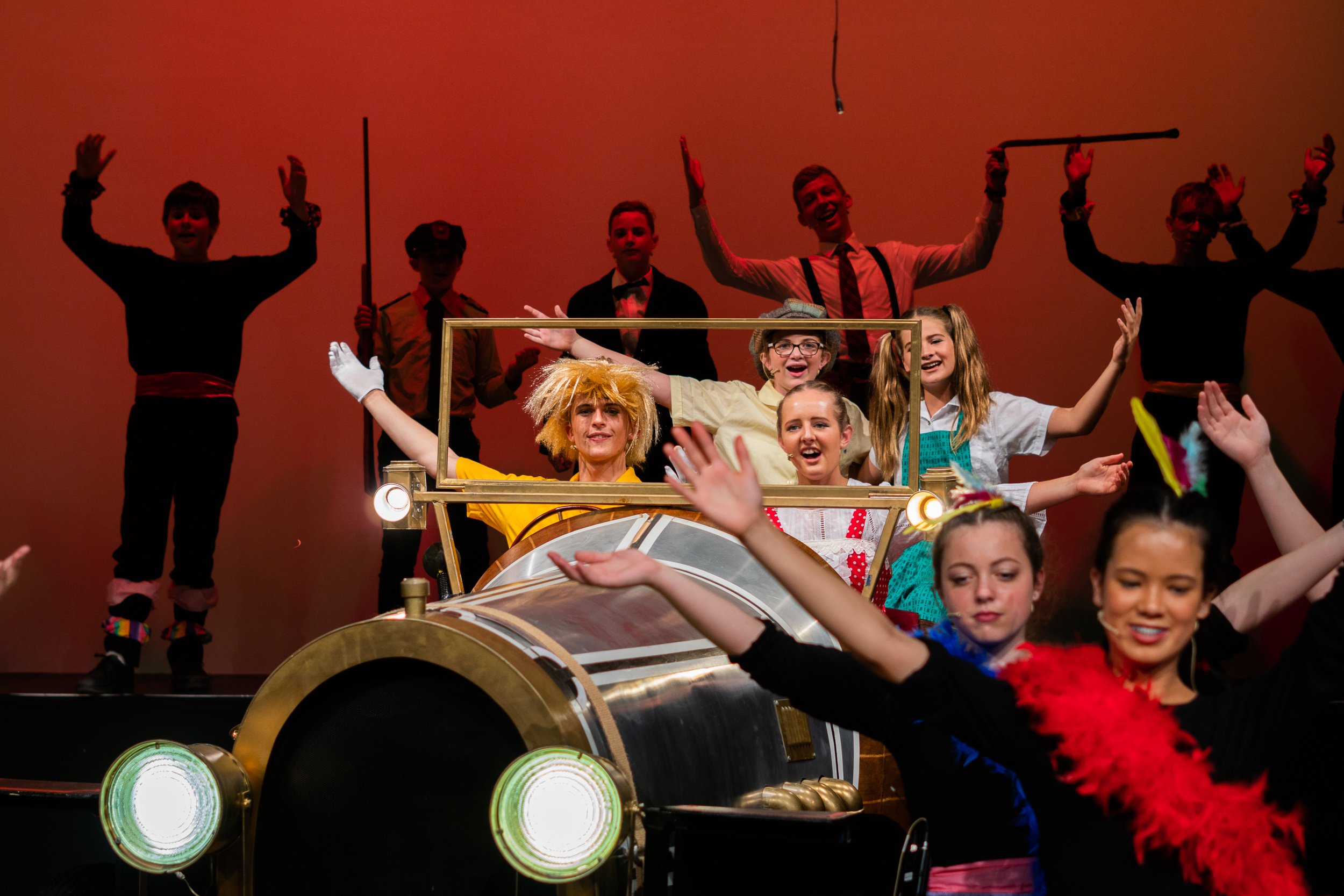 Children performing on stage with a vintage car prop, dressed in colorful costumes, with some kids raising their hands and others smiling, under warm stage lighting.