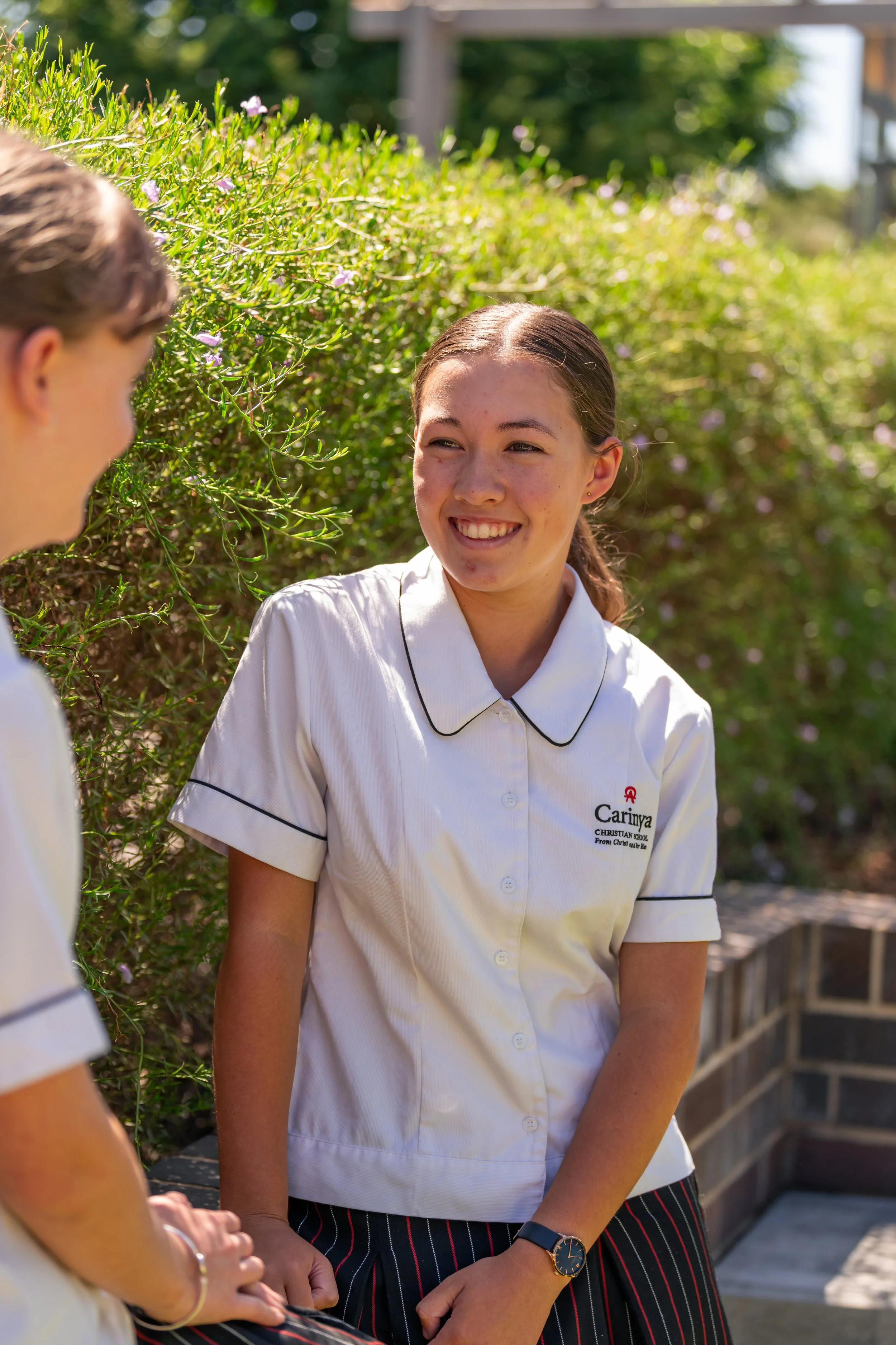 A girl in a white school uniform smiling and talking outdoors with another girl, with green bushes and outdoor furniture in the background.