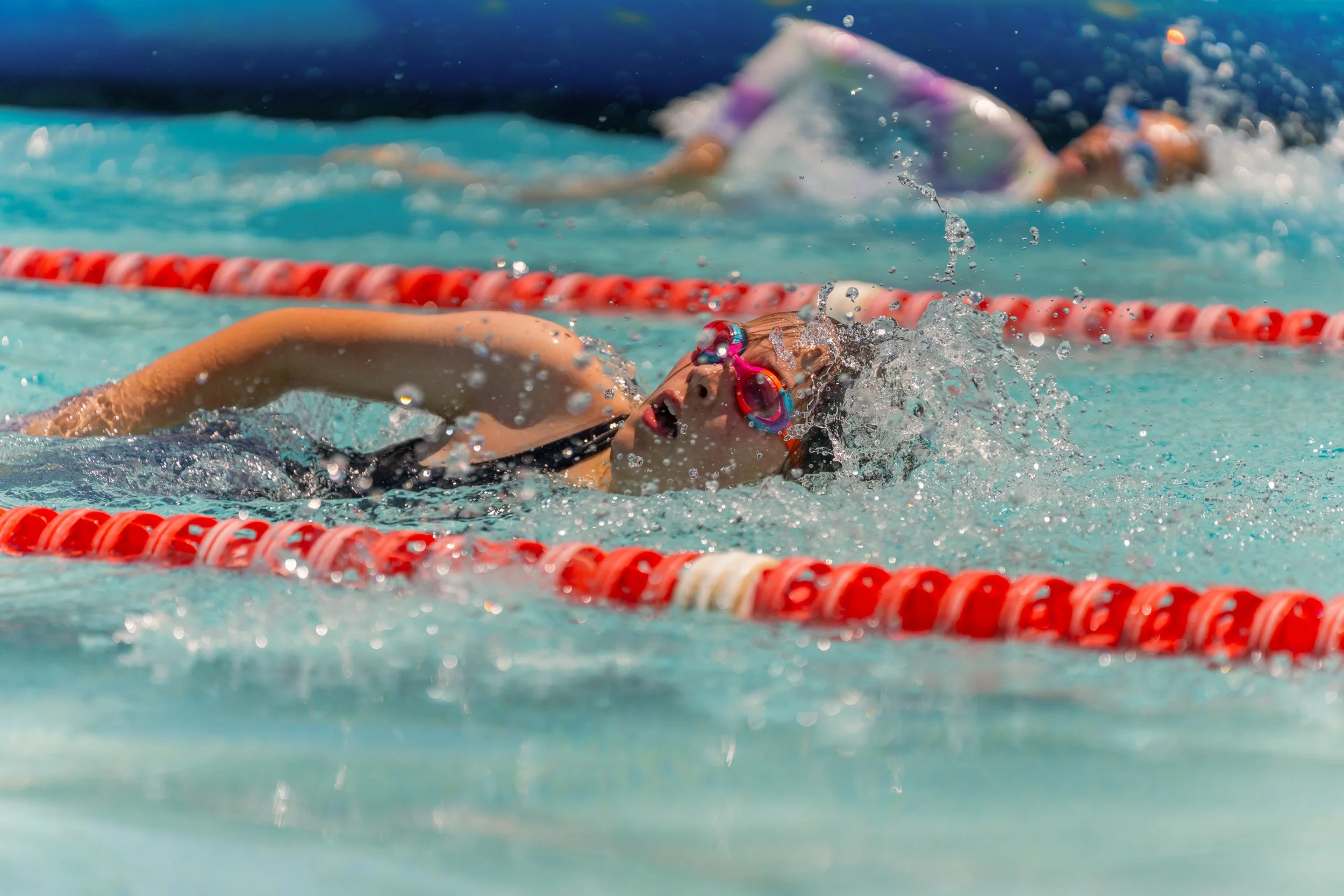 Swimmer in goggles and swimsuit swimming in a pool with red lane dividers, water splashing around.
