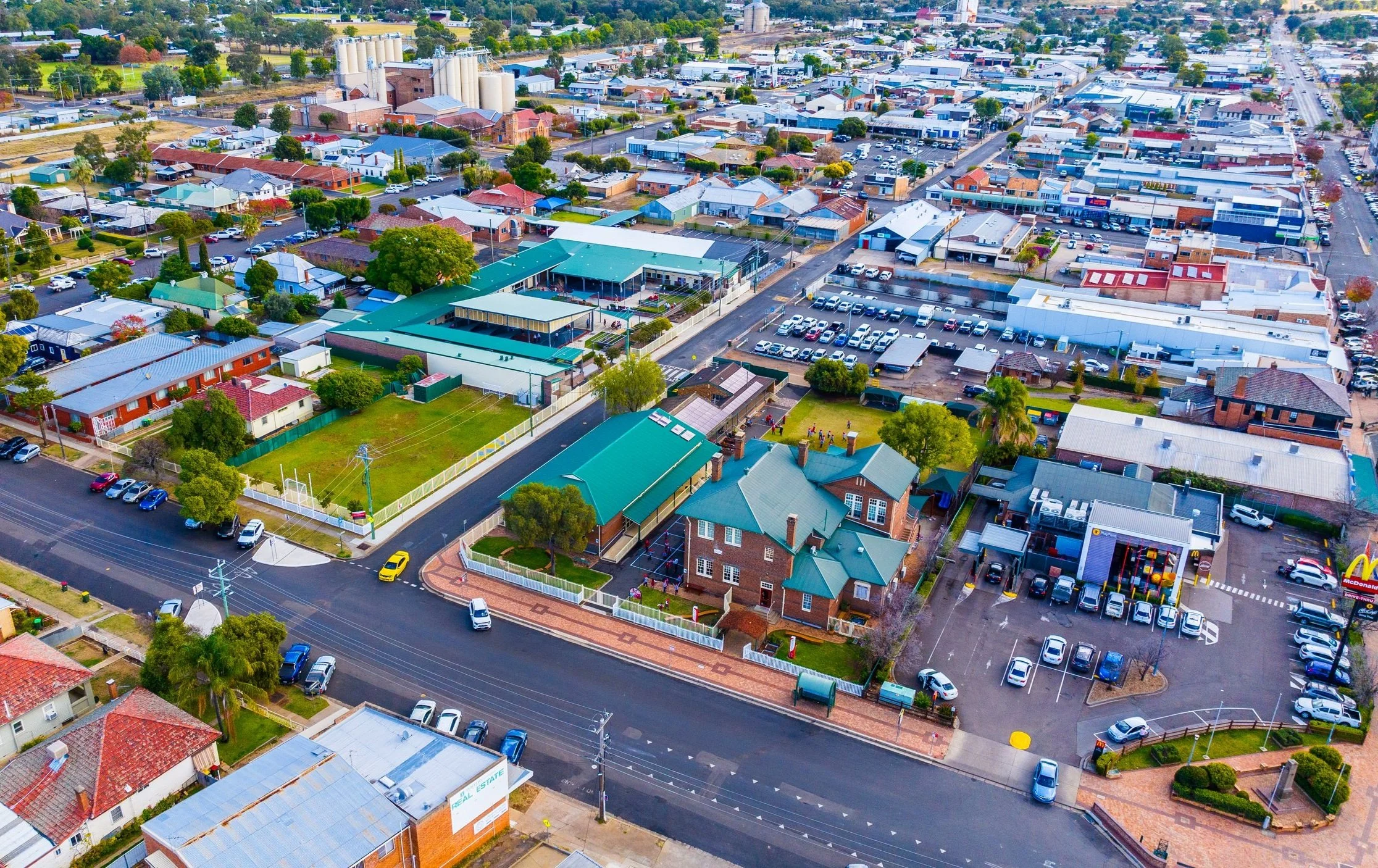 Aerial view of Carinya Christian School, Gunnedah, and surrounding residential houses, commercial buildings, parking lots, and streets. A School Building with a green roof and a schoolyard are visible.