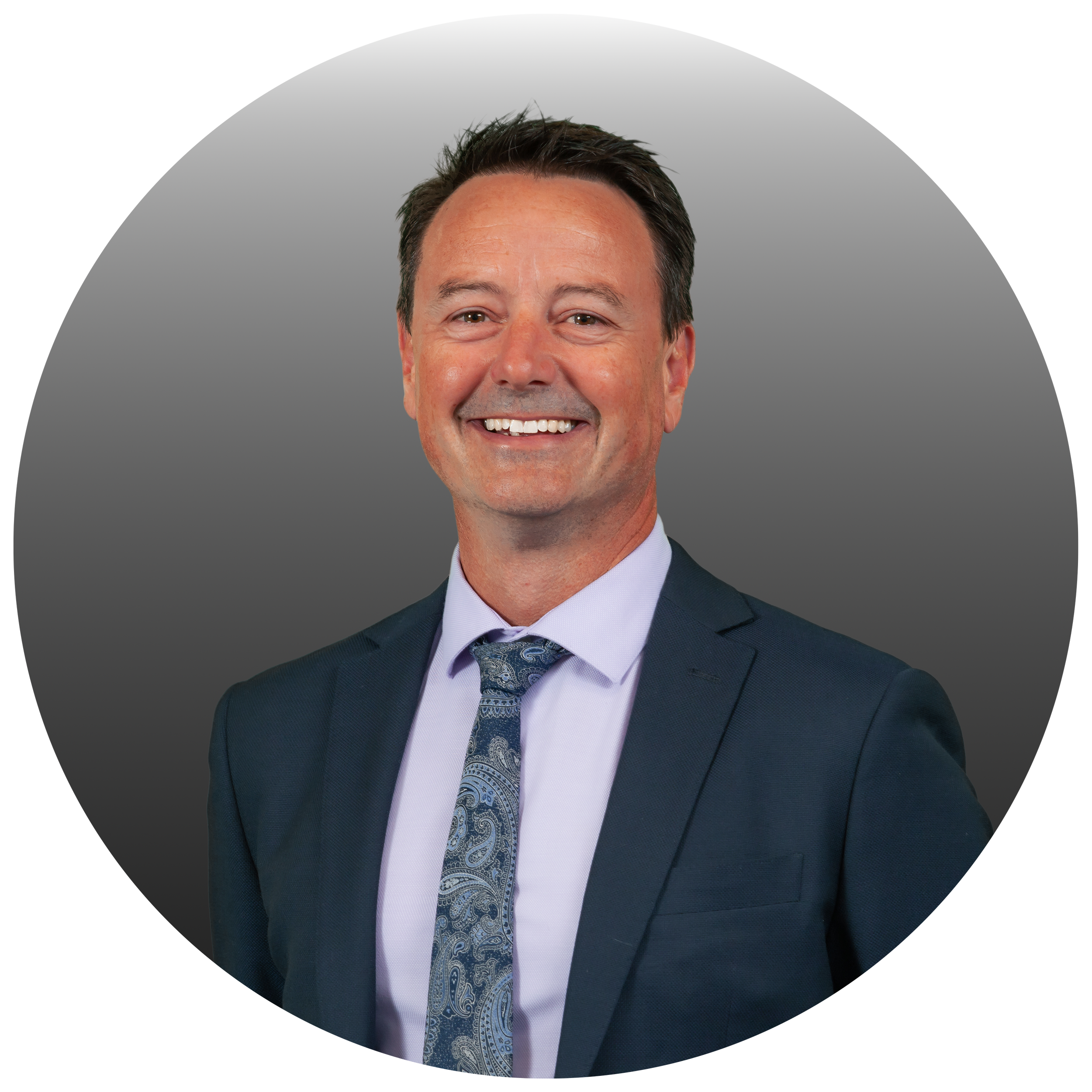 A smiling man in a dark suit, light blue shirt, and patterned tie, posing against a gray background.